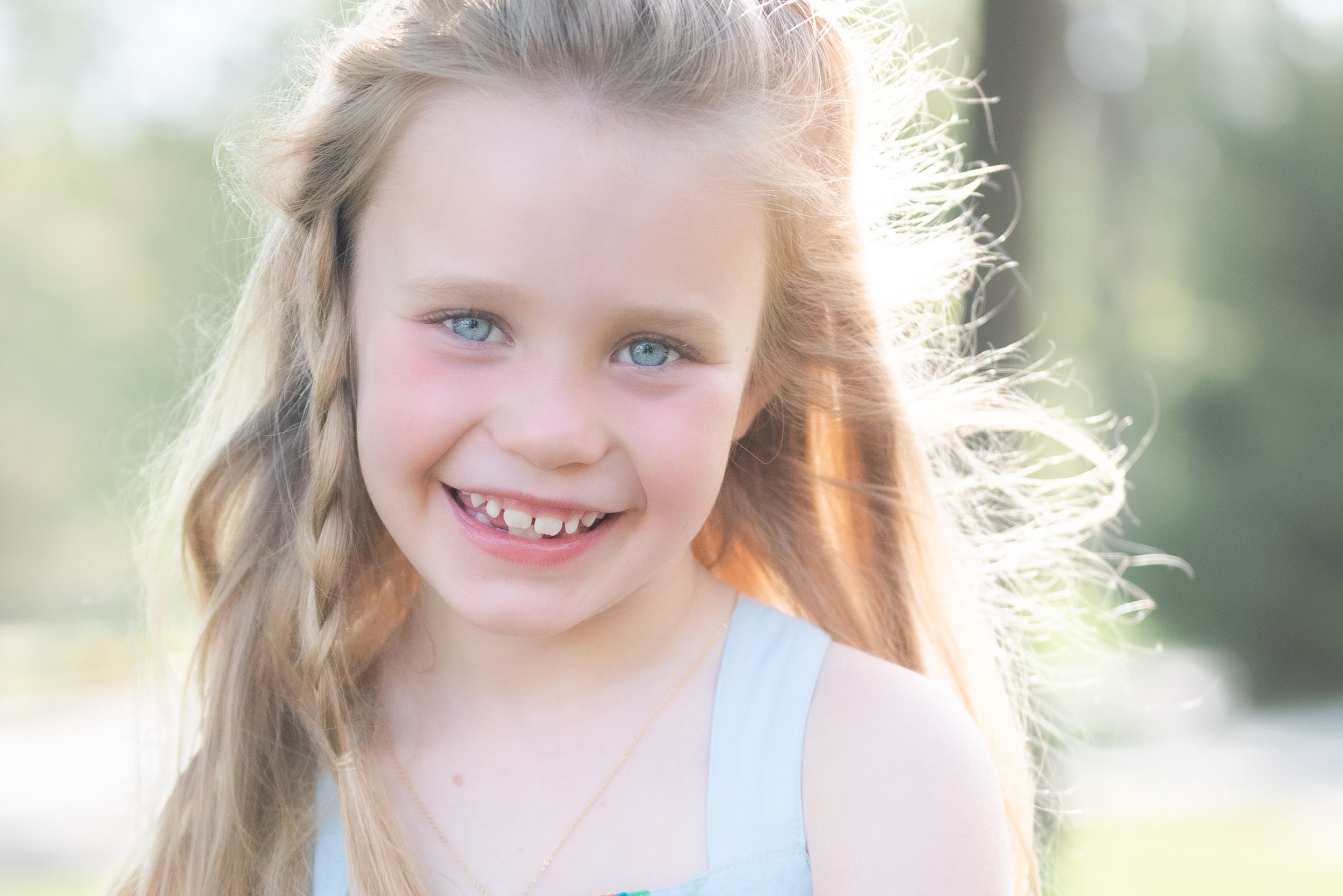 Smiling young girl with blue eyes and long wavy hair wearing a light blue top outdoors in soft sunlight.