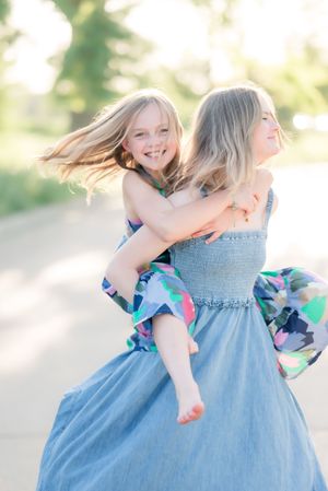 Two young girls in colorful dresses laugh joyfully as one gives the other a piggyback ride outdoors.