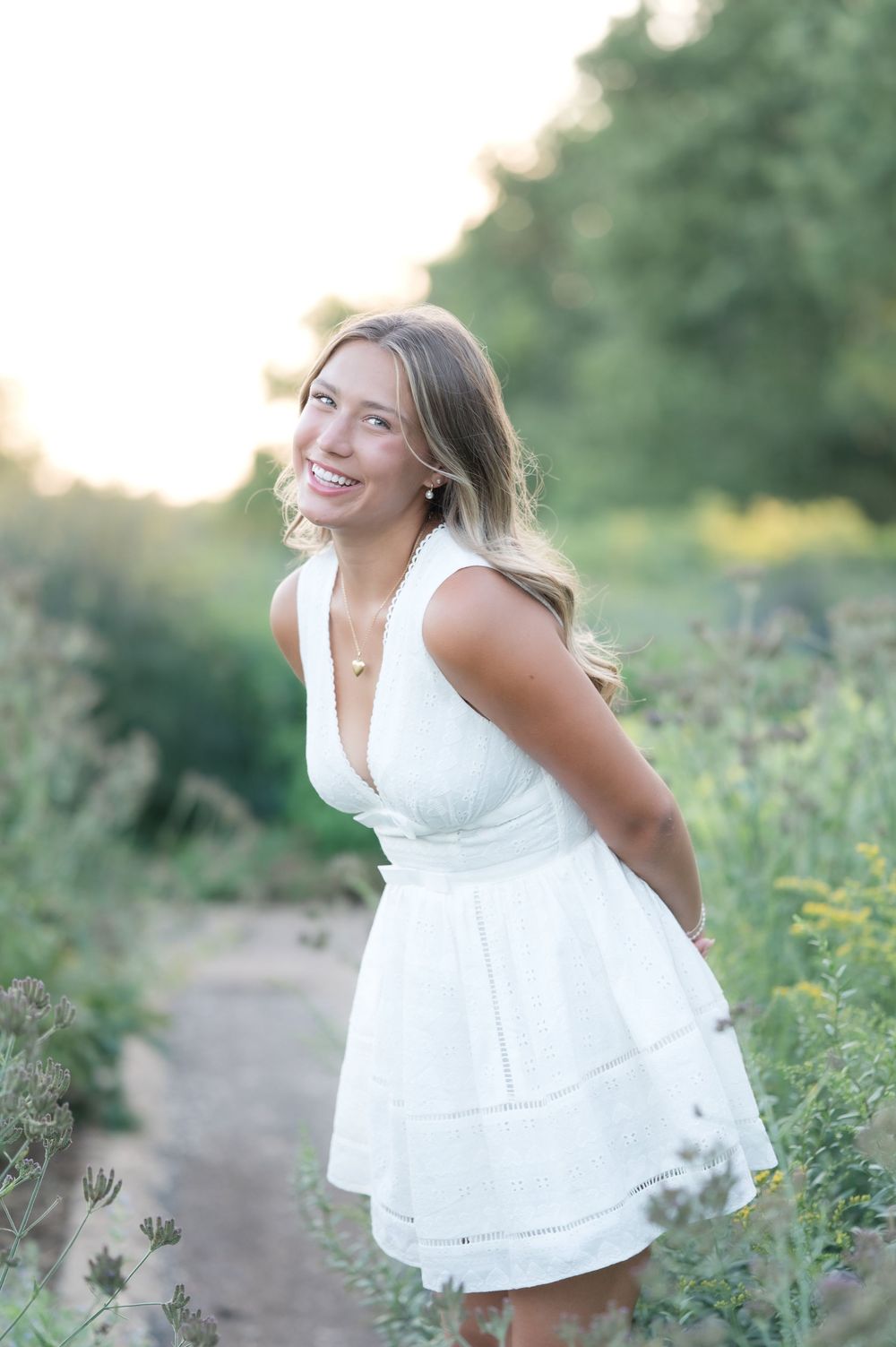 Young woman in white dress smiling over shoulder in sunlit wildflower field during golden hour.