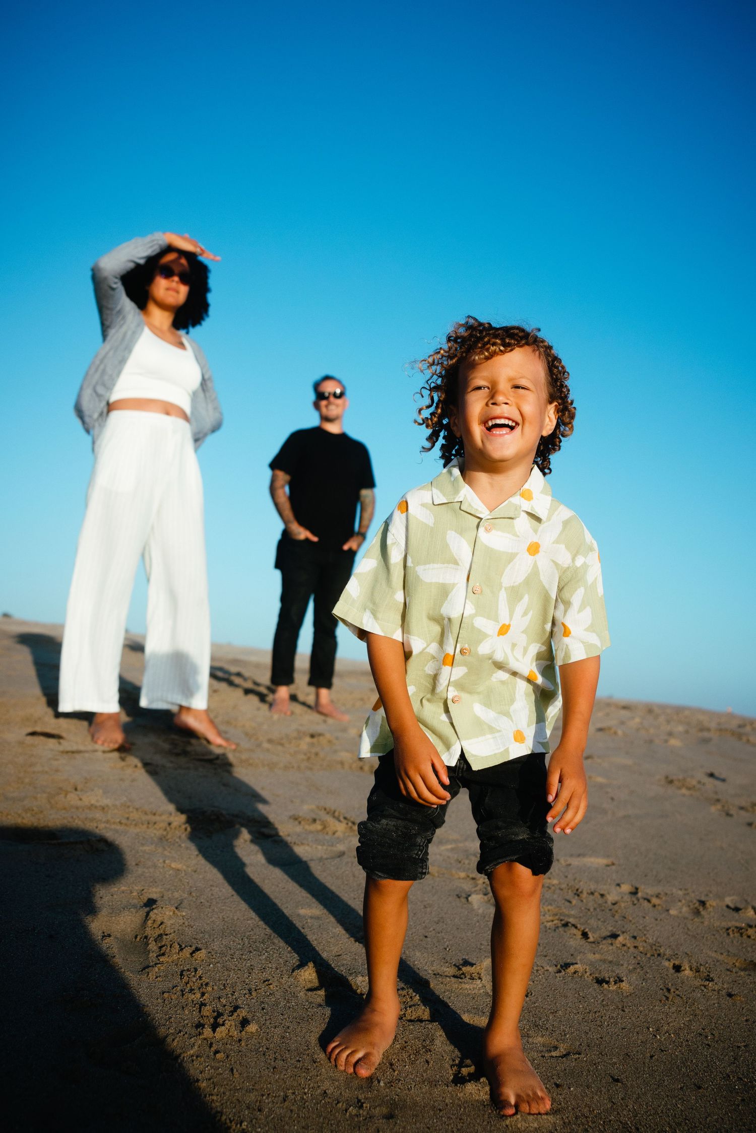 A joyful child laughs on a sunny beach while two adults stand in the background against a clear blue sky.