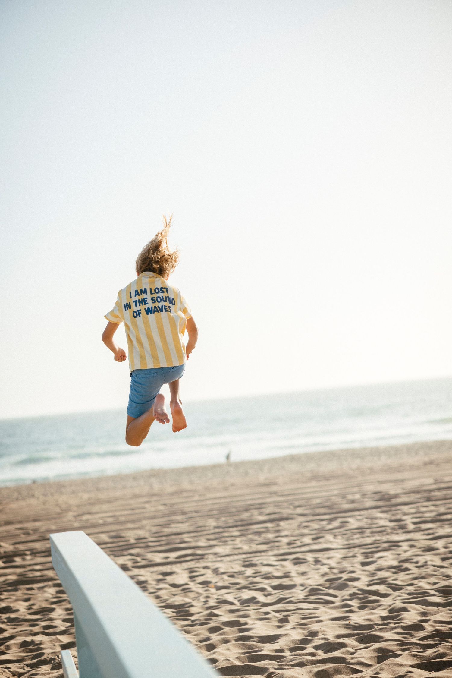 Someone wearing a t-shirt and shorts jumps energetically on a sunny beach next to a white barrier.