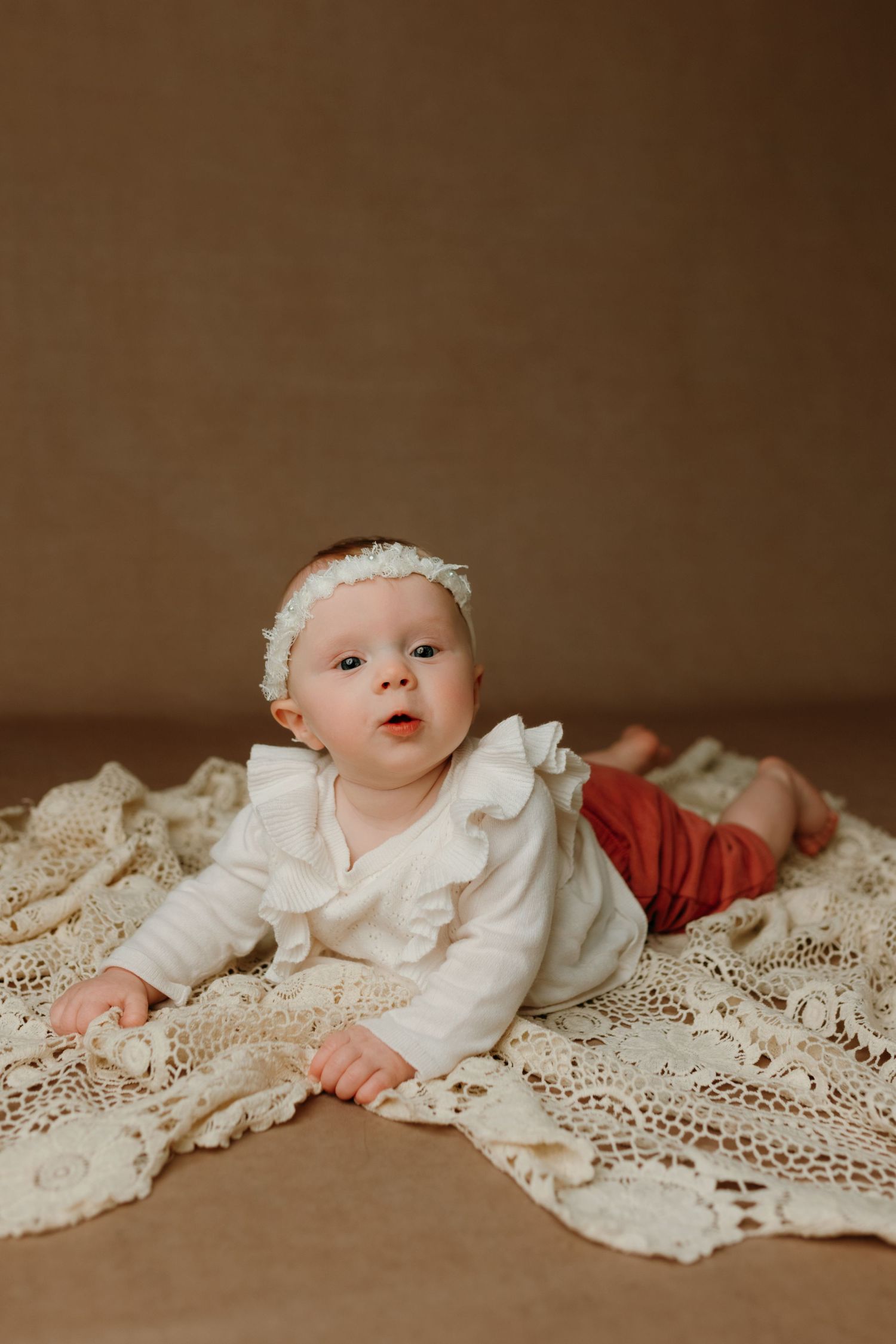 Baby wearing white ruffled dress lying on vintage lace blanket against brown backdrop during portrait session.