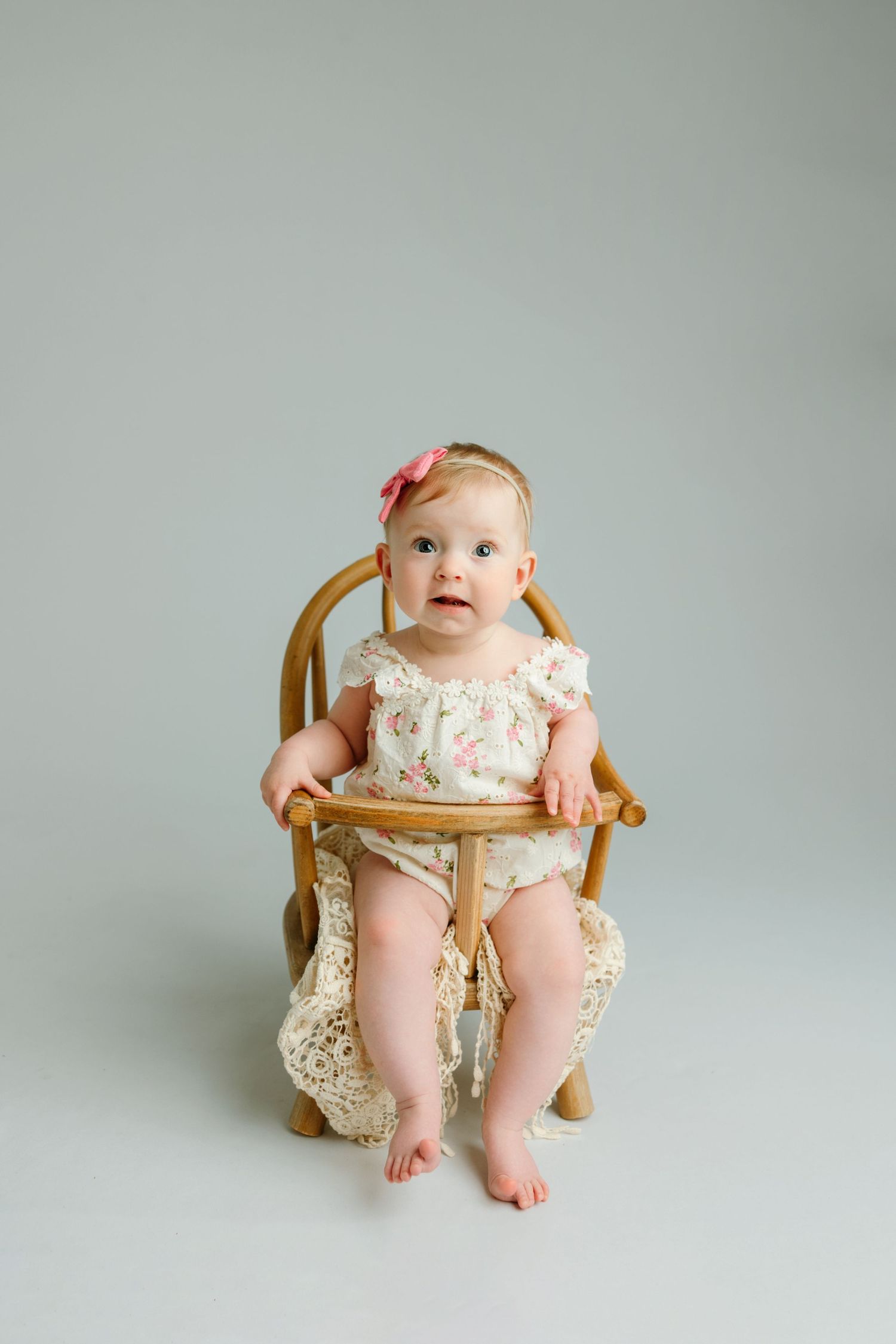 Baby girl in floral romper and pink bow headband sitting on wooden chair against gray background at the baby photo studio in Victoria BC.
