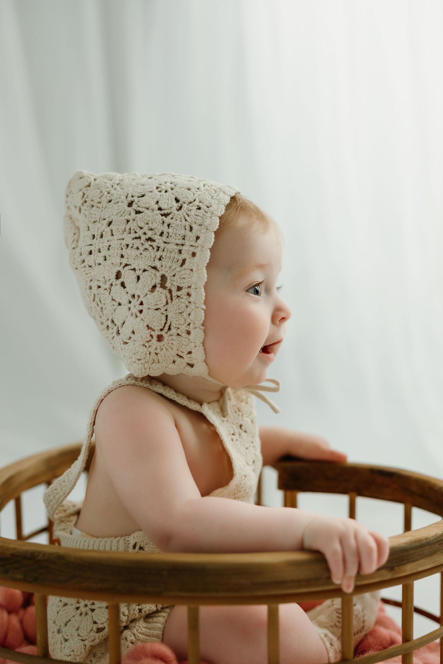 Baby in crochet bonnet and romper sitting in a wooden bassinet against a white backdrop at the baby photo studio in Victoria BC.