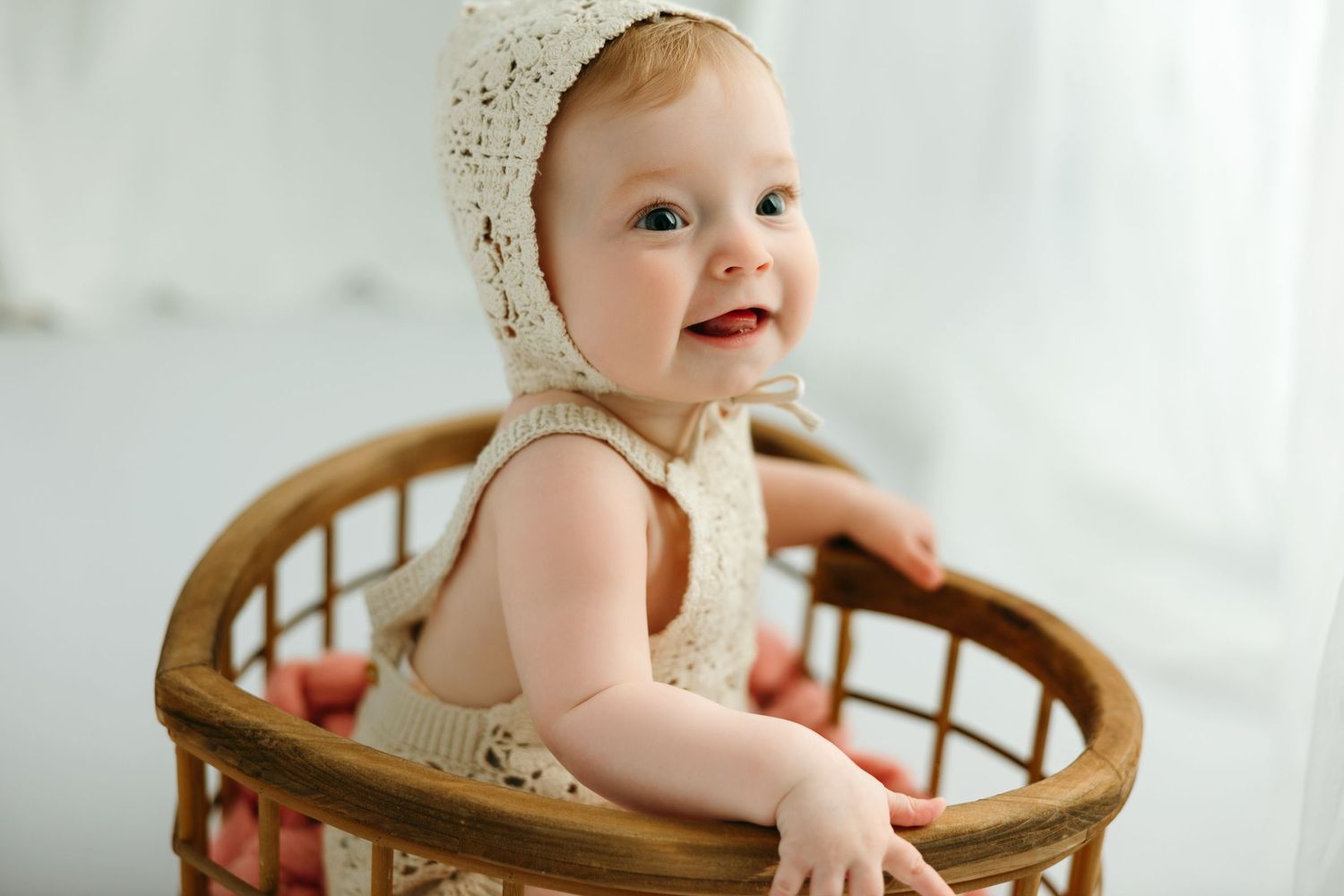 Smiling baby in cream lace bonnet and romper sitting in a wicker basket in a bright white baby photo studio in Victoria BC.