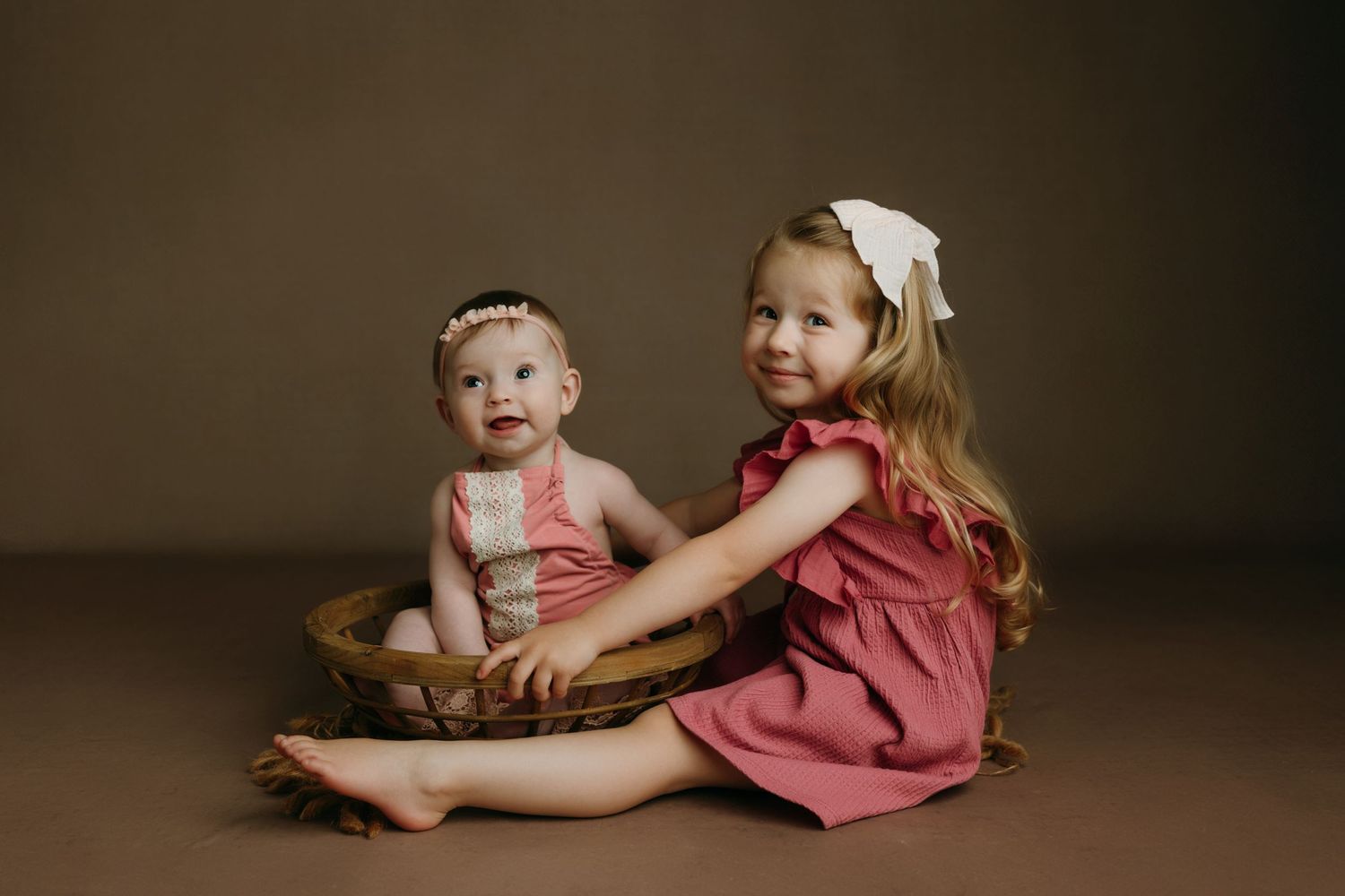 baby sisters pose together, older girl in pink dress with bow hugs baby sibling sitting in a wooden bowl basket at the baby studio yyj.