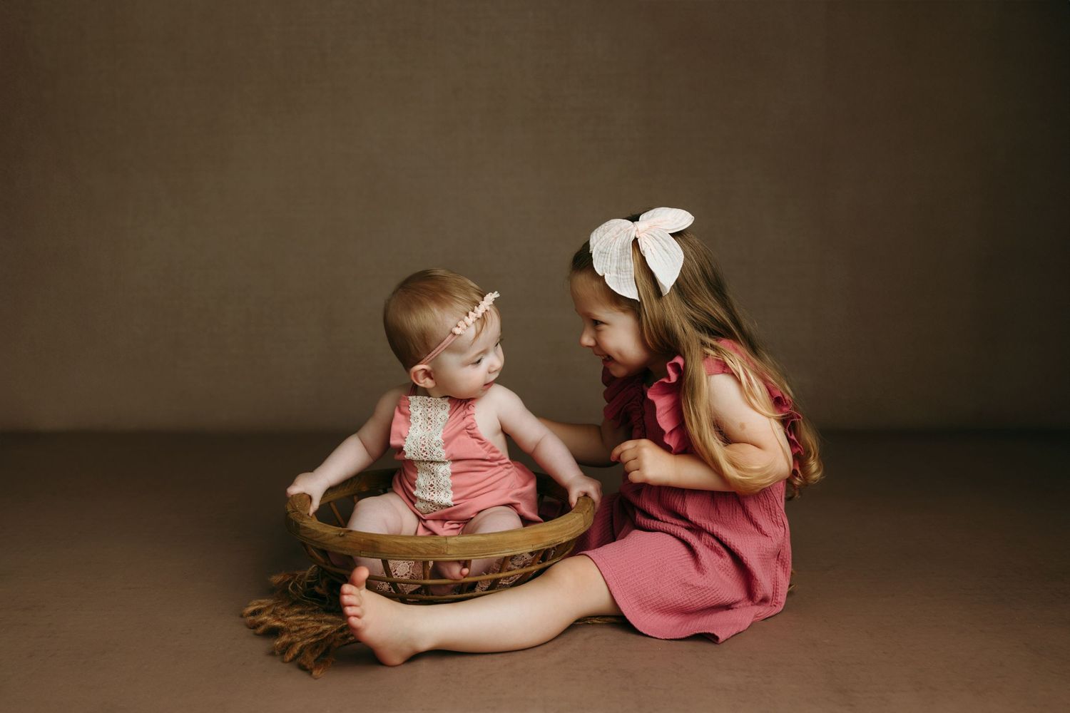Two young sisters in pink outfits share a sweet moment, older girl smiling at baby sitting in a woven bowl at the baby photo studio yyj.