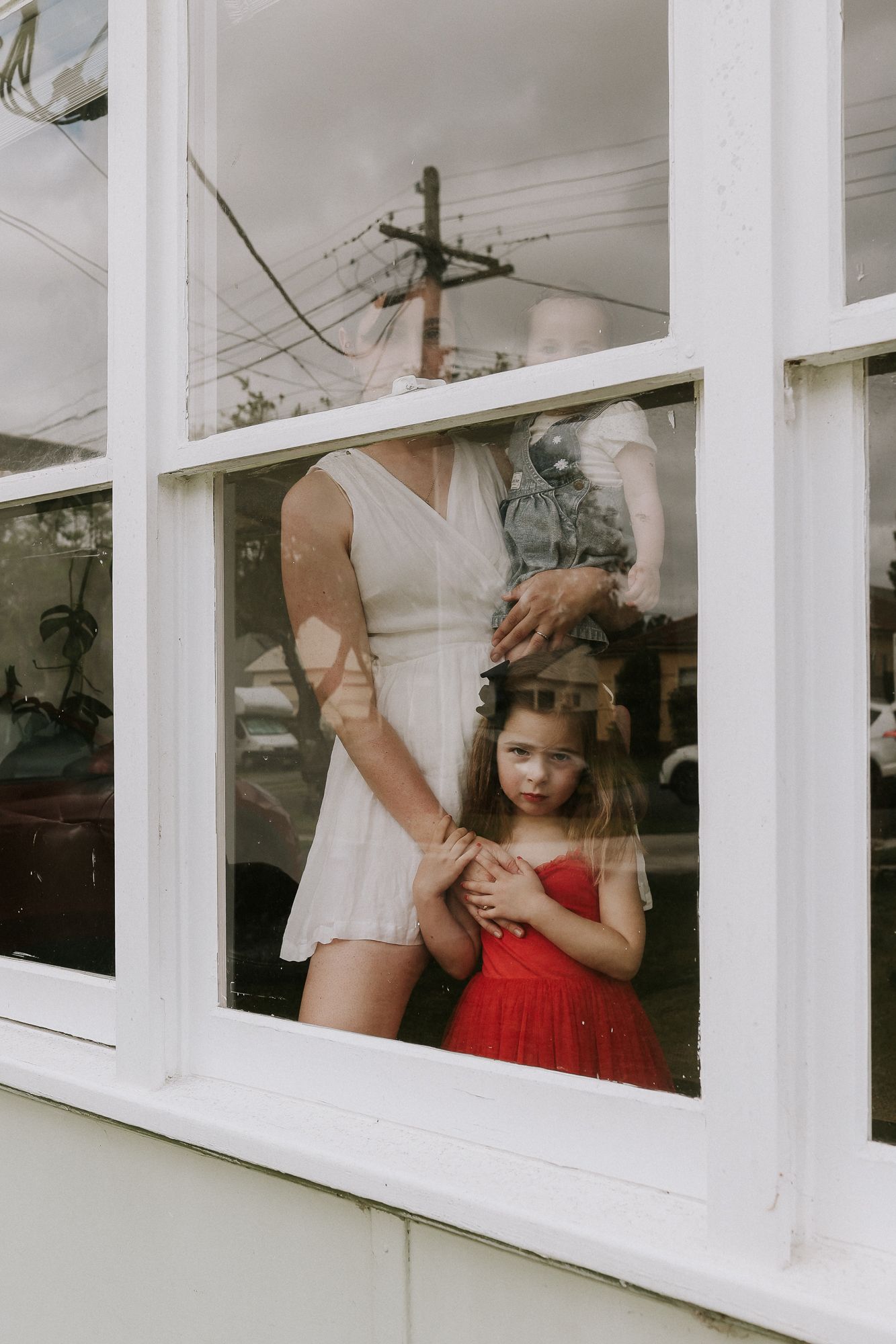 A young girl in a red dress stands with a woman in white at a window with reflections visible outside.