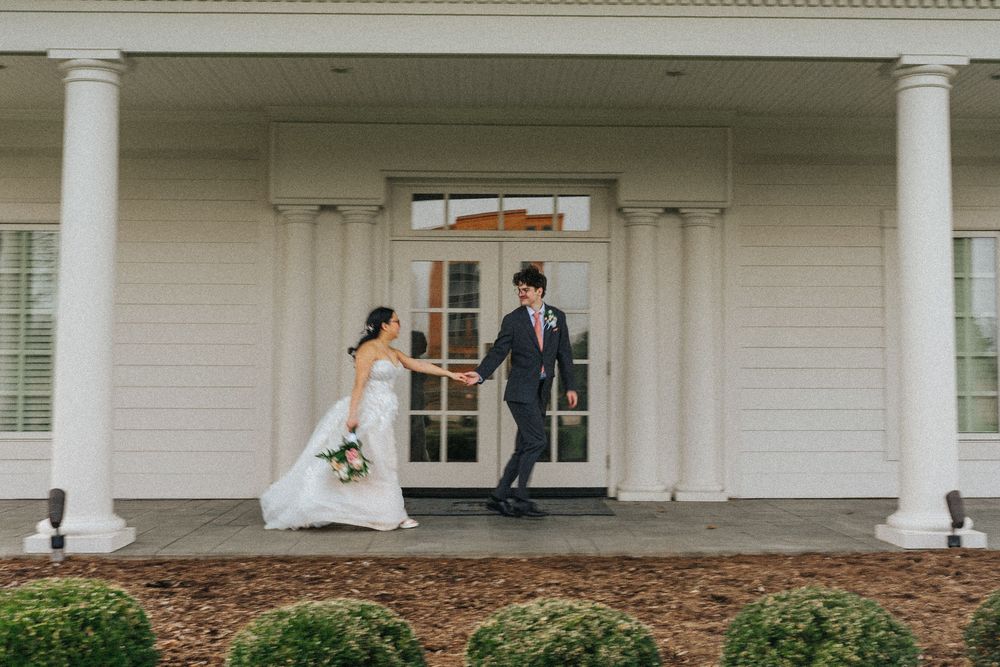 Bride and groom hold hands on a white columned venue porch, bride in white gown holding bouquet, groom in navy suit.