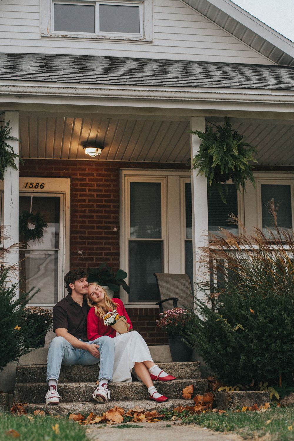 Couple sitting on front porch steps of a brick house numbered 1586, sharing a tender moment amid autumn leaves.