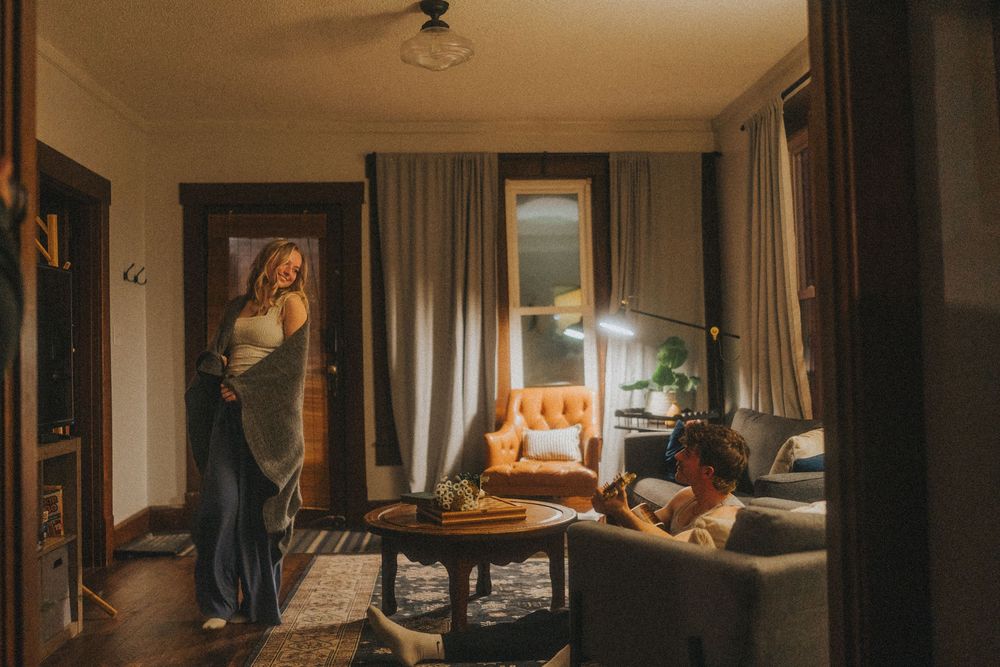 Woman standing in doorway while another person relaxes on sofa in warmly lit cozy living room.