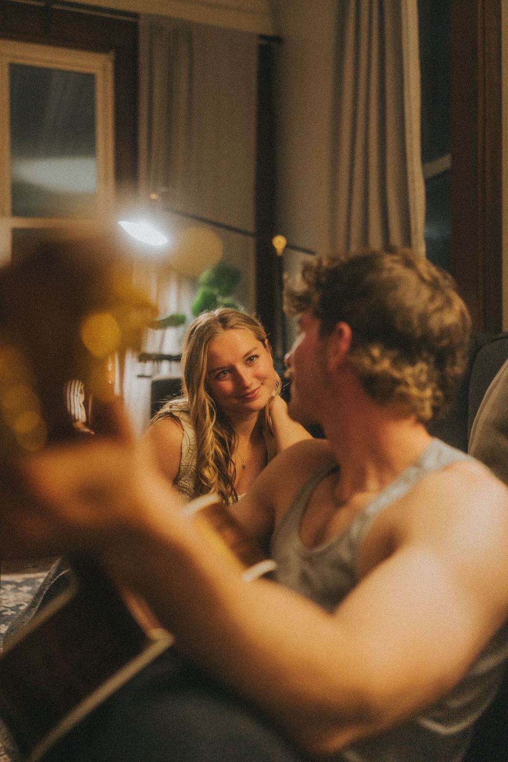 A couple shares a romantic moment indoors, woman smiling warmly as man plays guitar in cozy warm-lit room.