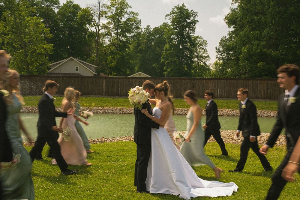 Bride and groom share a kiss outdoors at a wedding ceremony while the bridal party walks past on a green lawn.
