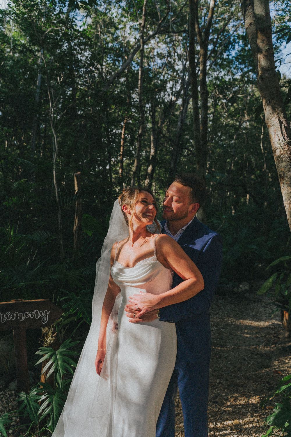 Bride and groom embrace in a lush forest setting, she wears a white gown with veil, he wears a navy suit.