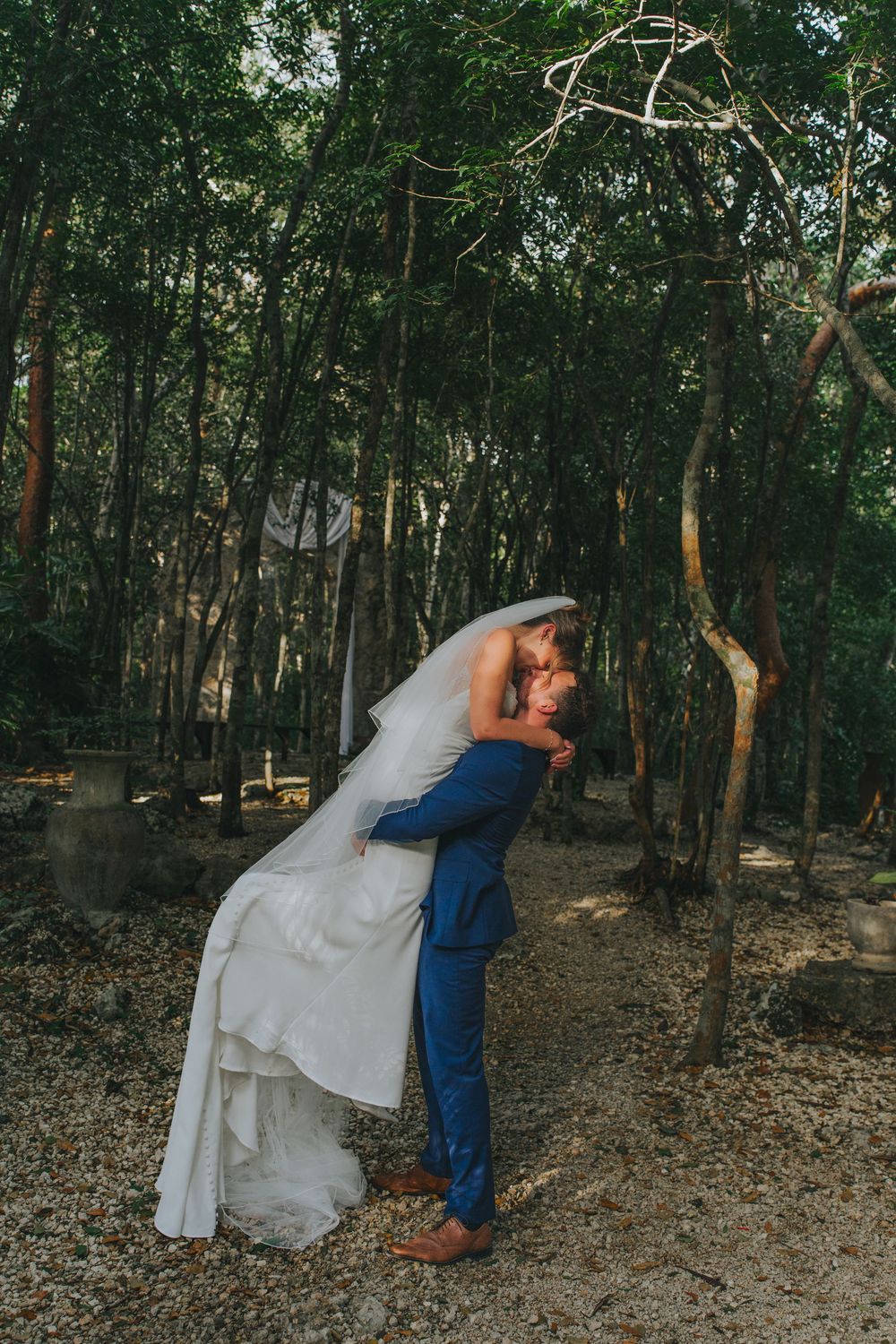Bride and groom share a romantic dip kiss surrounded by lush green trees in an outdoor forest wedding setting.