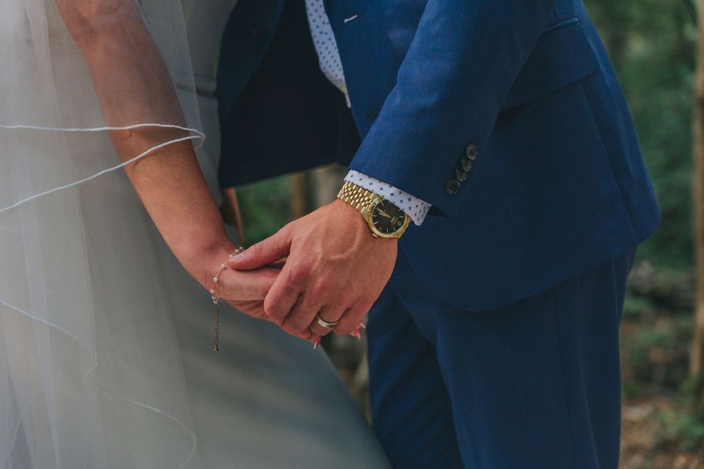 Bride and groom holding hands, groom wearing blue suit and gold watch, bride in white wedding dress.