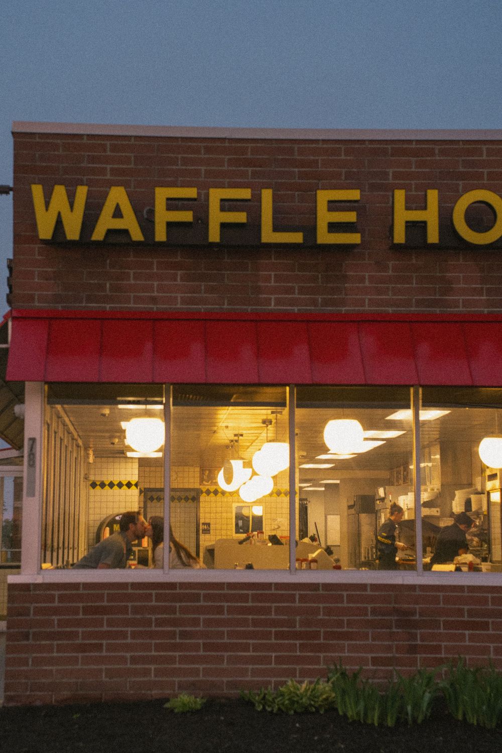 Waffle House restaurant exterior at dusk with glowing yellow sign and red awning, customers visible through large windows.