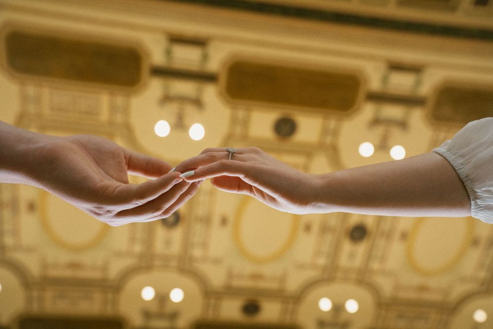 Two hands gently touching fingertips in an elegant golden ballroom, with a ring visible on one finger.