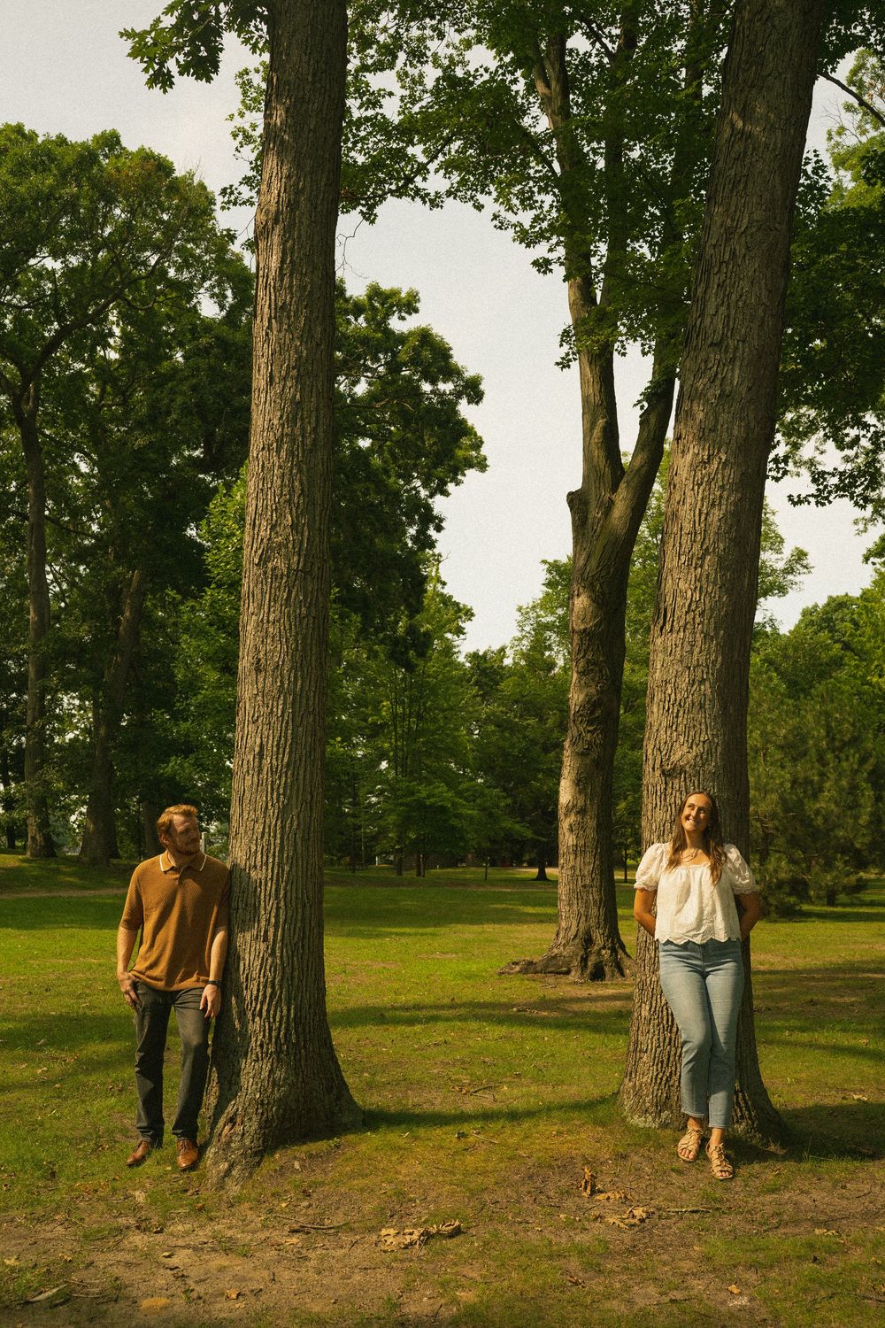 Couple standing apart by tall trees in a lush green park, man in brown shirt and woman in white top and jeans.