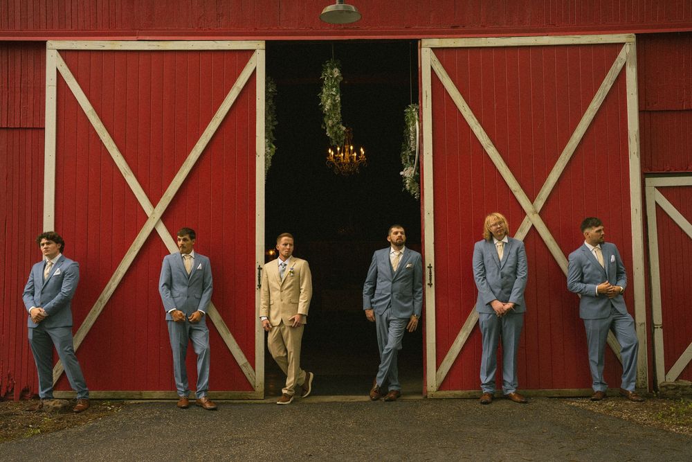 Groomsmen in blue suits stand before a rustic red barn with white trim doors at a wedding venue.