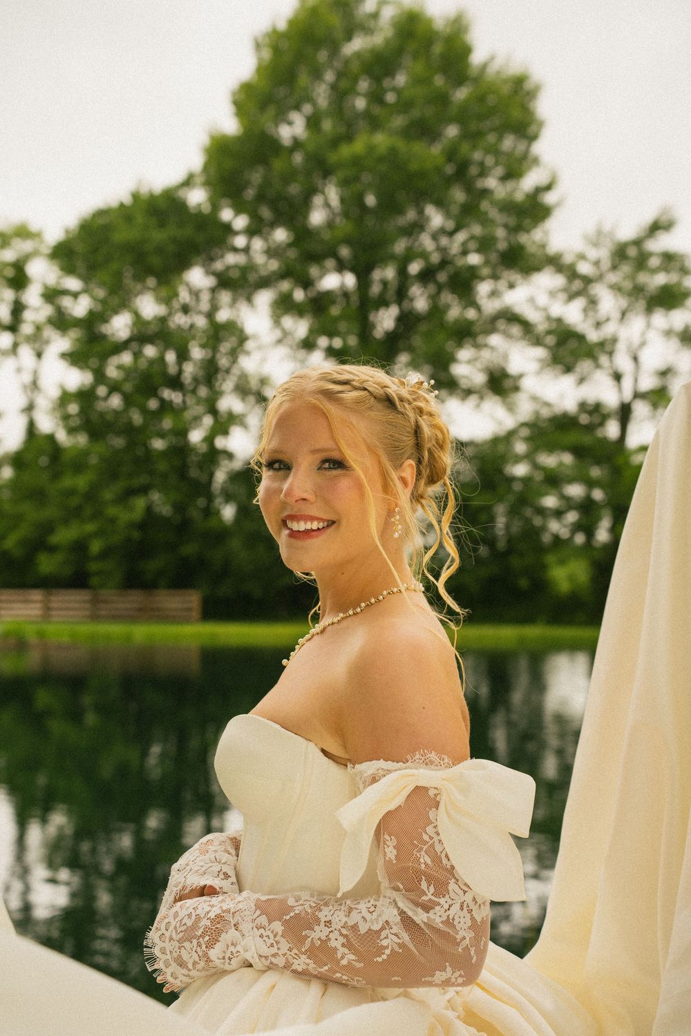 Smiling blonde bride in off-shoulder lace wedding gown with pearl necklace poses outdoors near a pond.