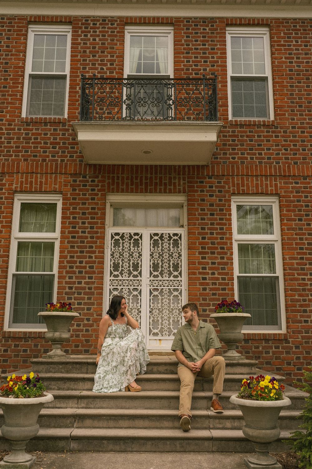 Couple sitting on steps of elegant brick mansion with ornate white door, balcony, and colorful flower planters.