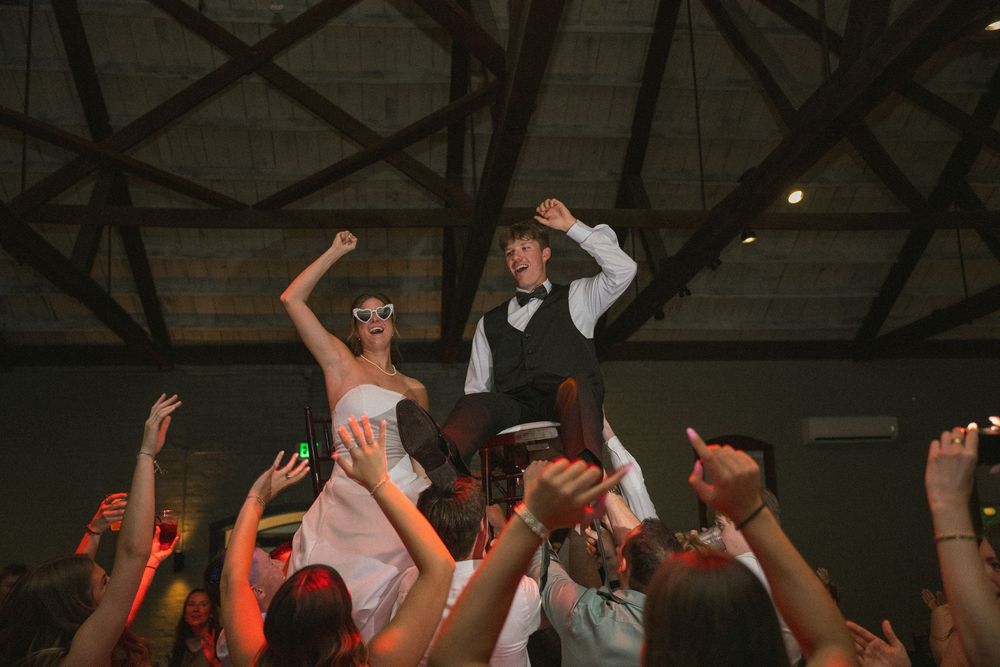 Bride and groom lifted by guests on dance floor at lively wedding reception celebration.