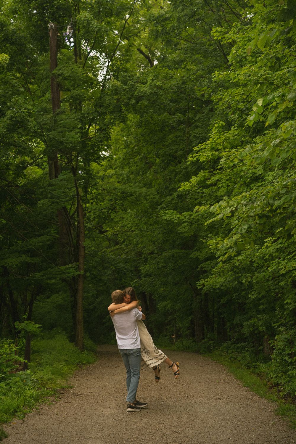 Couple embracing on a tree-lined path, man lifting woman in a romantic outdoor setting surrounded by lush green forest.