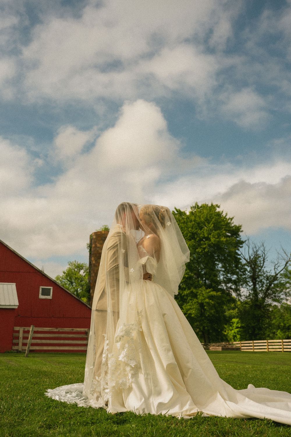 Two brides in white gowns and veils share a romantic moment on a green lawn beside a red barn under a cloudy sky.