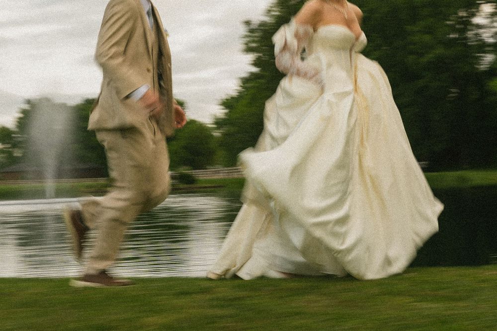 Bride in white gown and groom in tan suit running on green grass near a pond at an outdoor wedding.
