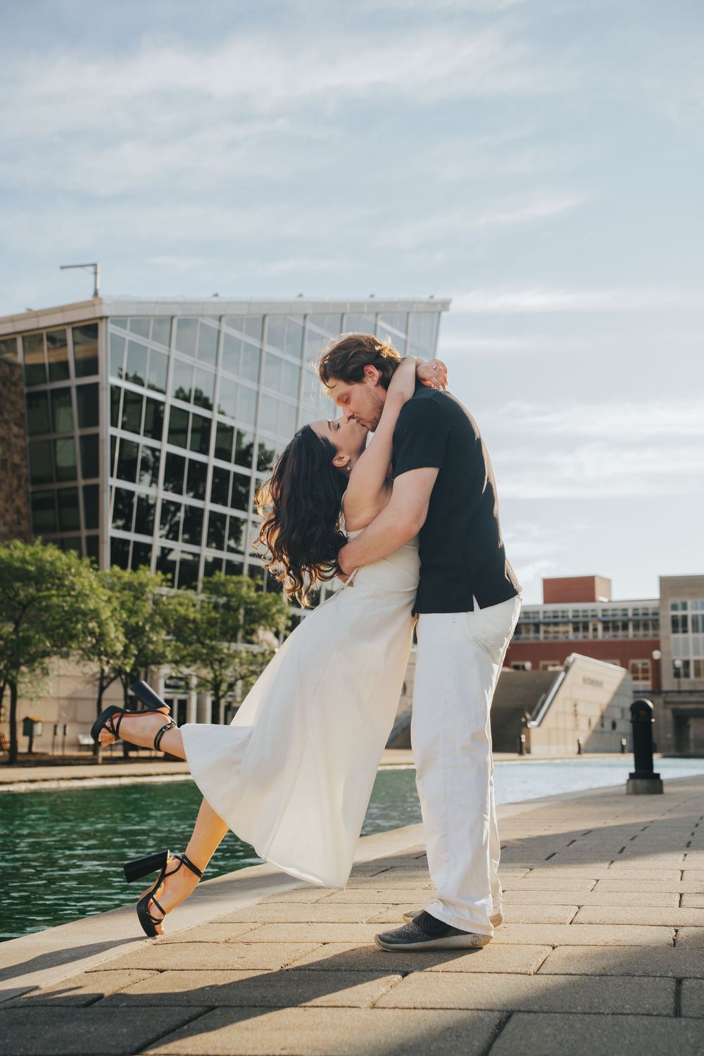 A romantic couple dancing in an embrace by a fountain with modern architecture in the background at sunset.