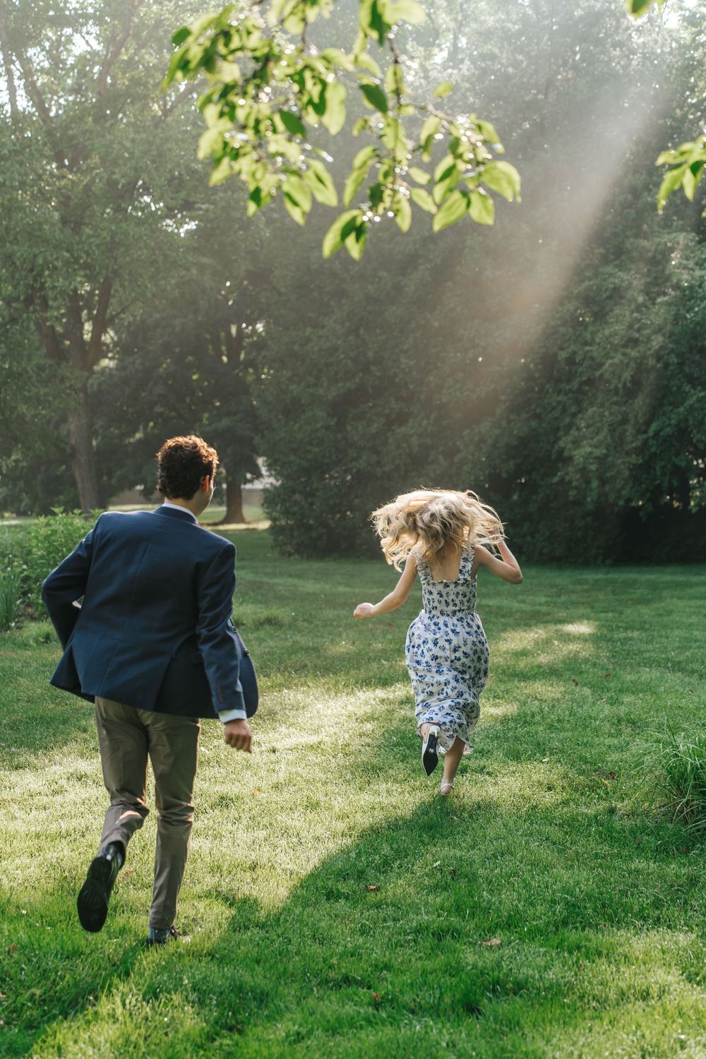Two people running playfully through a sunlit grassy field in a park during golden hour.