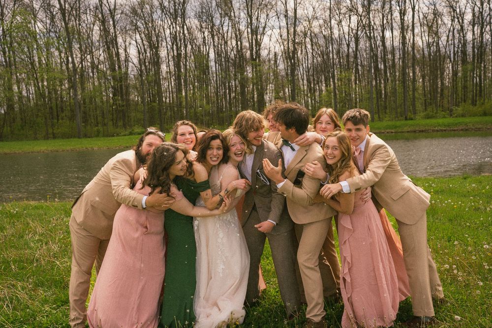 Wedding party in pink dresses and tan suits hugging bride and groom outdoors near a pond and trees.