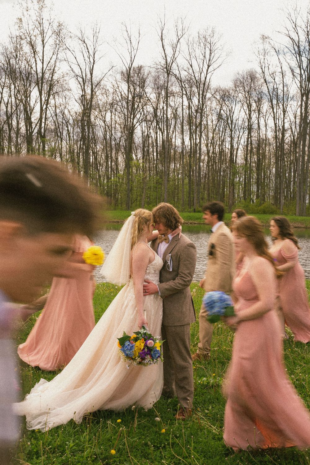 Wedding couple kissing surrounded by bridesmaids in pink dresses near a pond with bare trees backdrop.