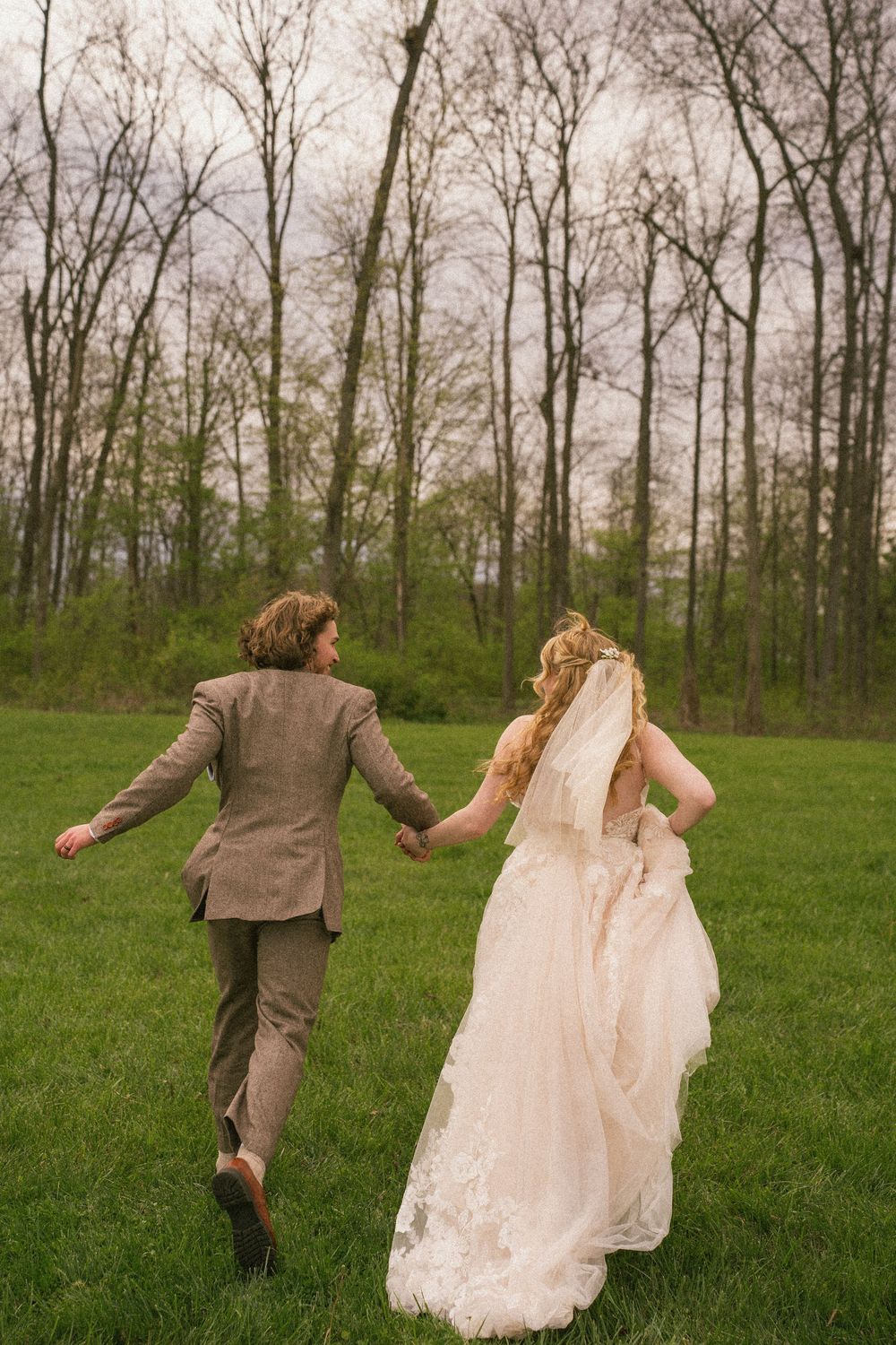 Bride and groom holding hands running through a green meadow with bare trees, bride in lace gown with veil.