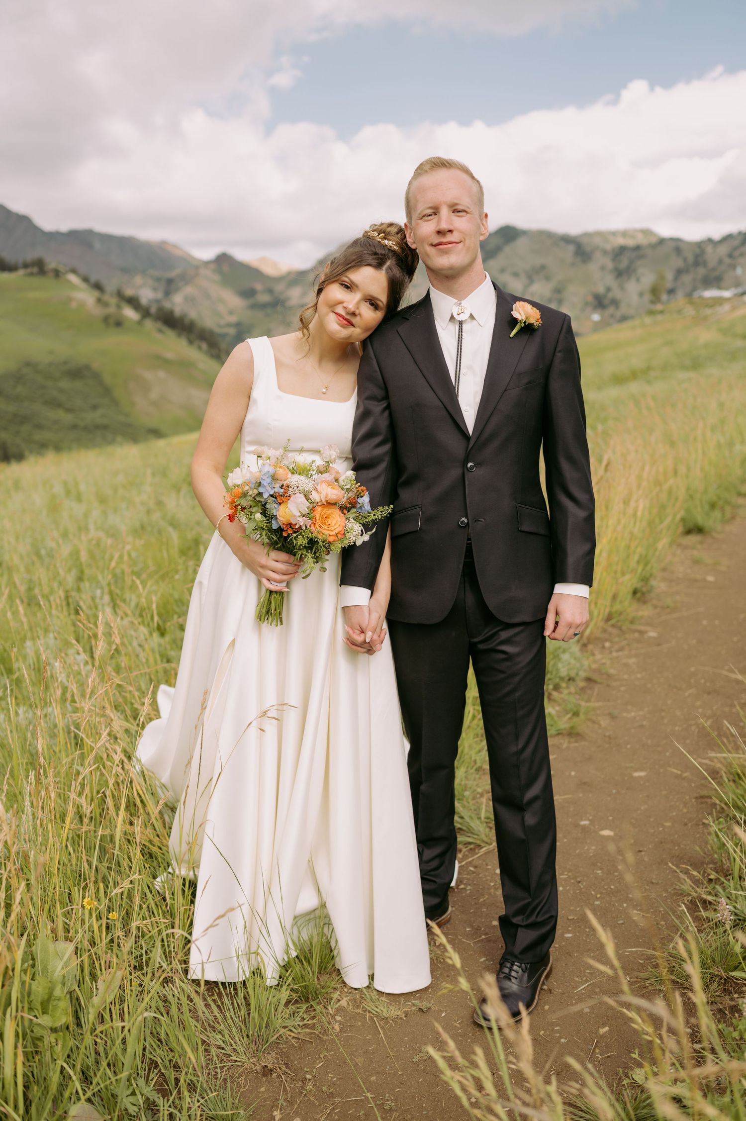 Happy newlyweds pose on mountain trail with colorful bouquet and scenic backdrop.