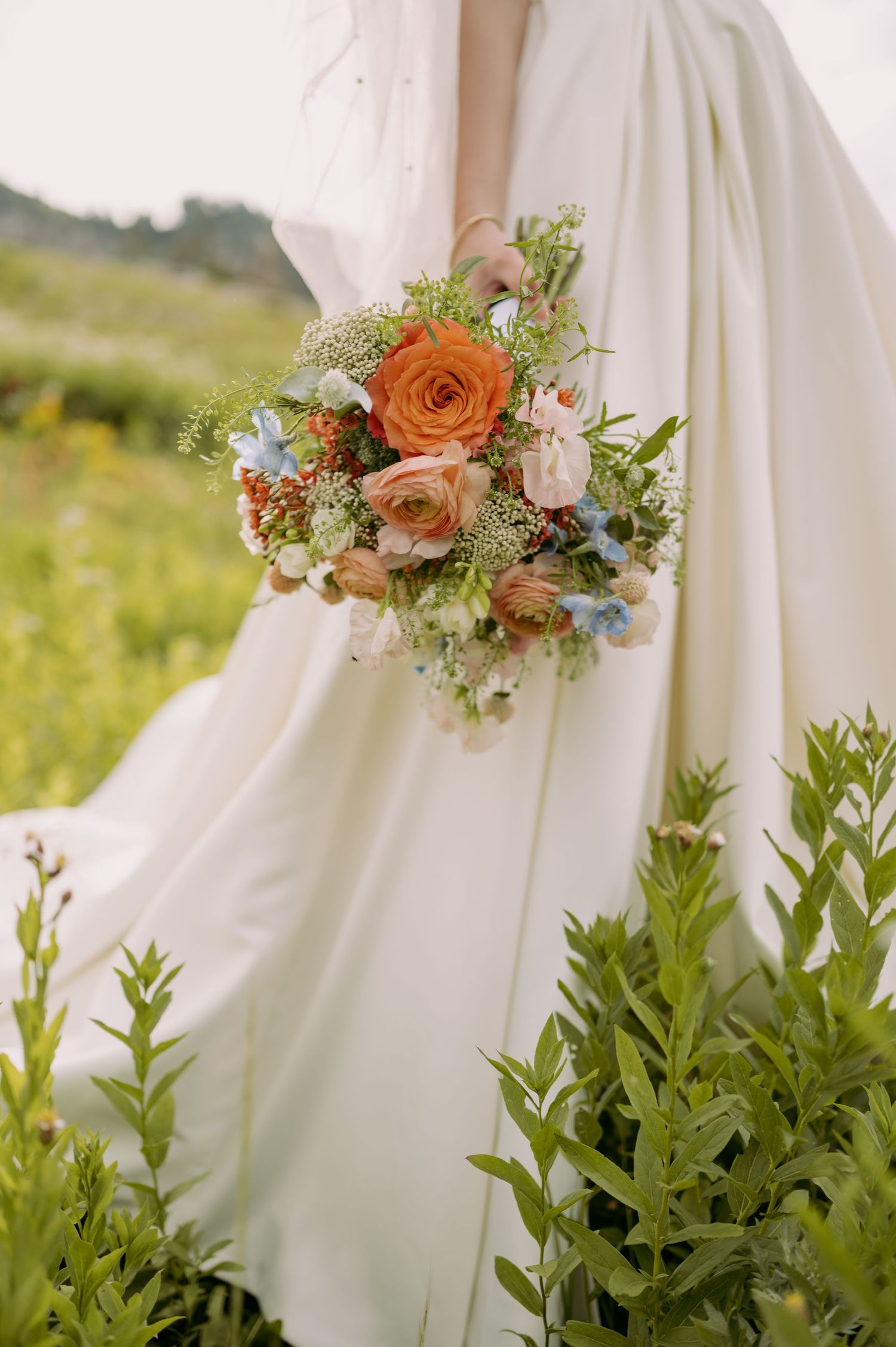 A romantic wedding bouquet with orange roses and wildflowers against a flowing white wedding dress in a meadow.