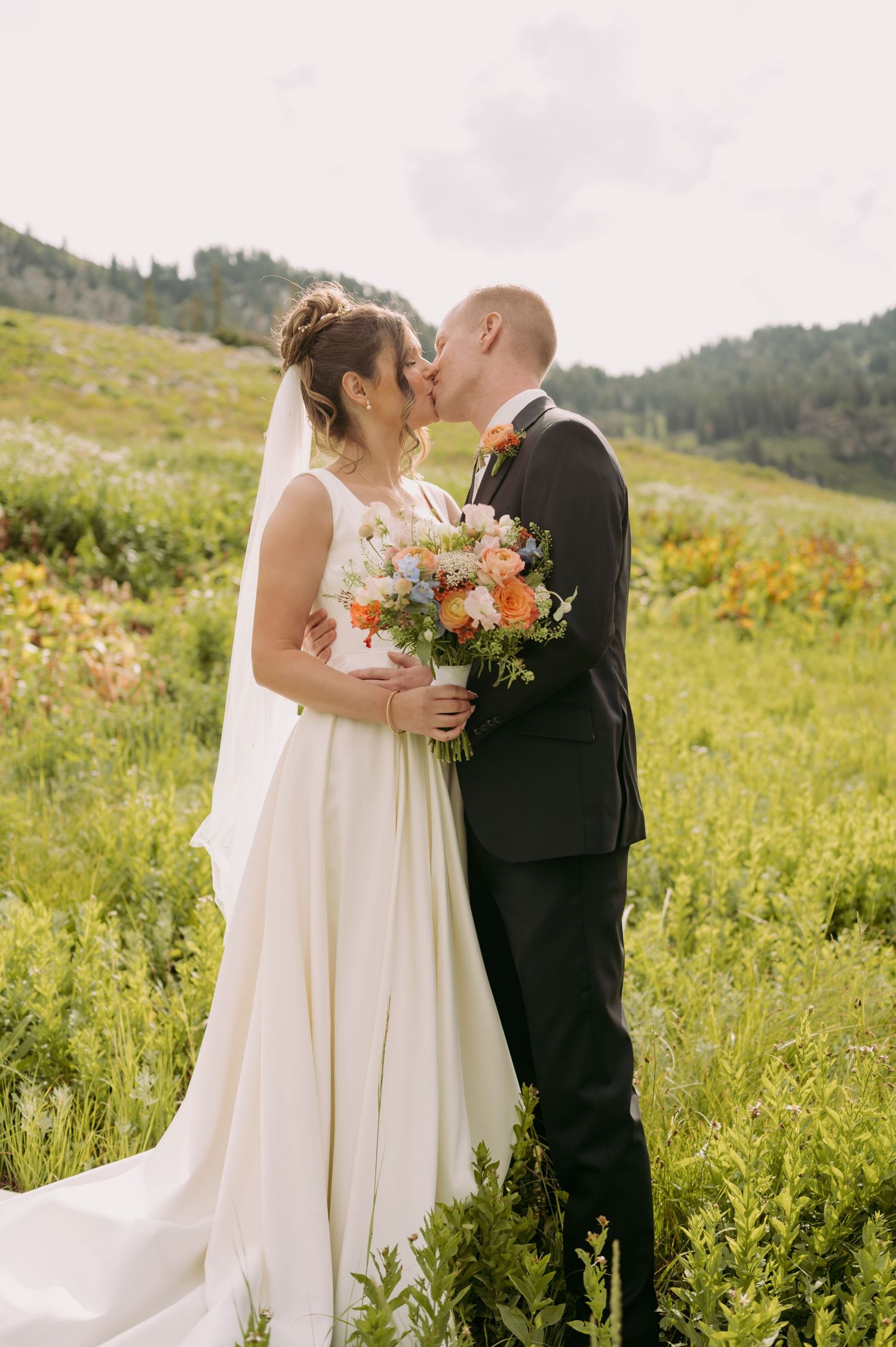A couple embraces in wedding attire against a mountainous backdrop with wildflowers.