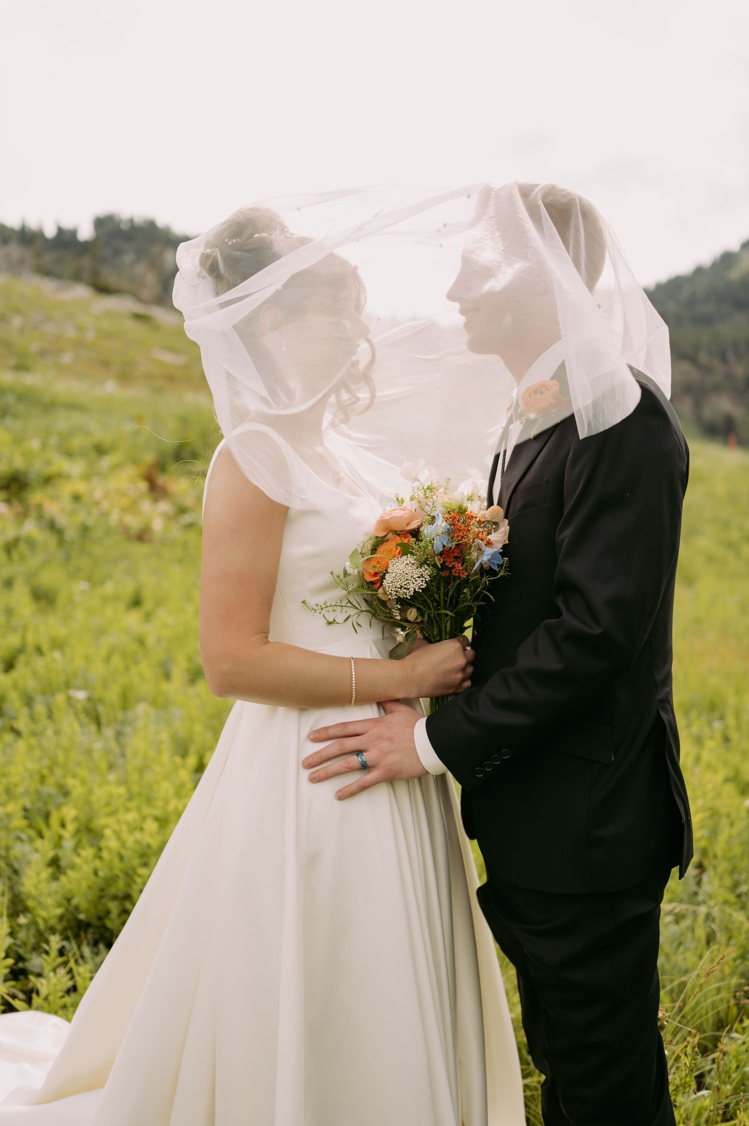 Wedding couple embraces under shared veil in mountain wildflower field.