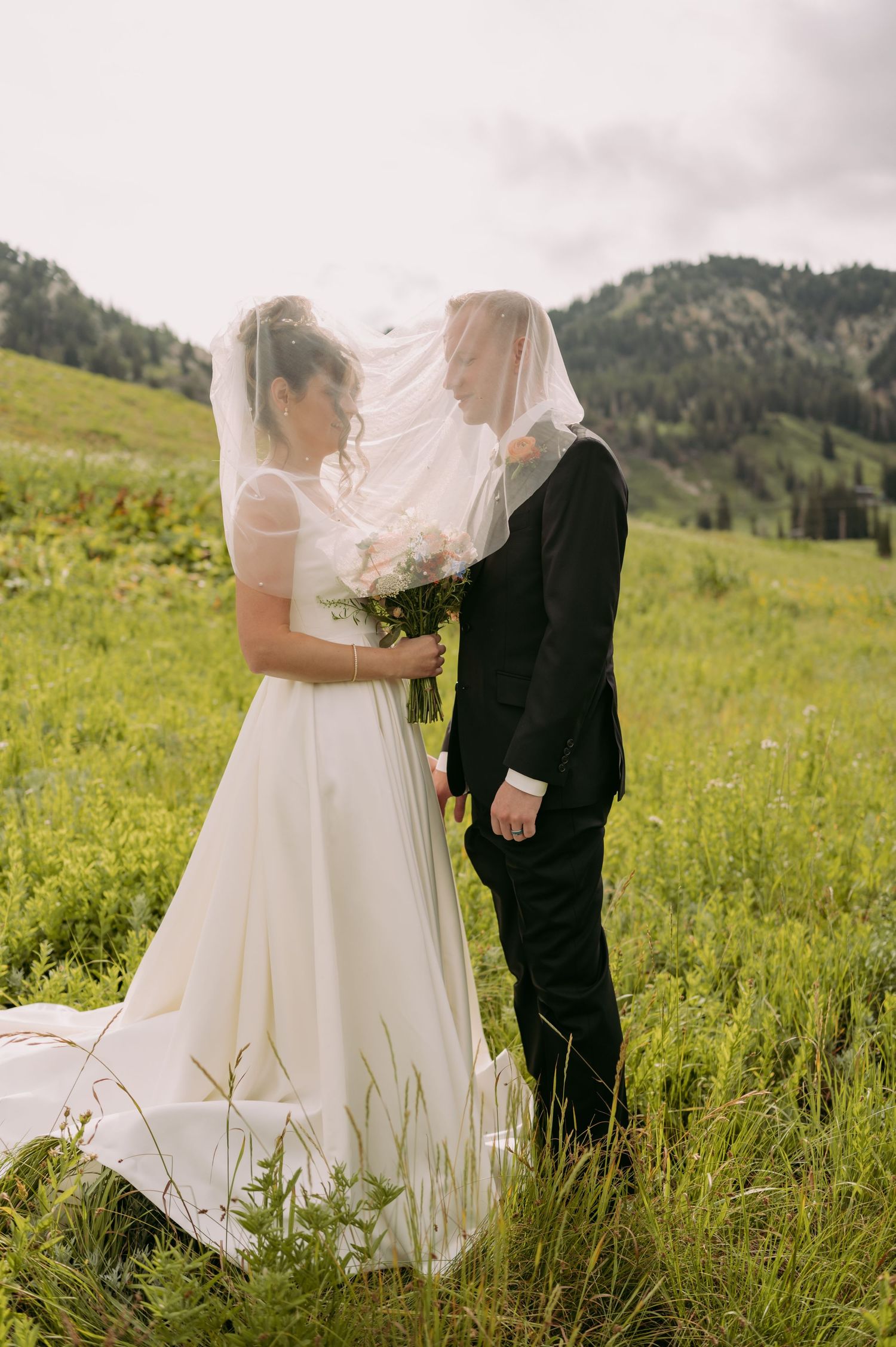 Newlyweds share intimate moment under flowing veil in mountain meadow.