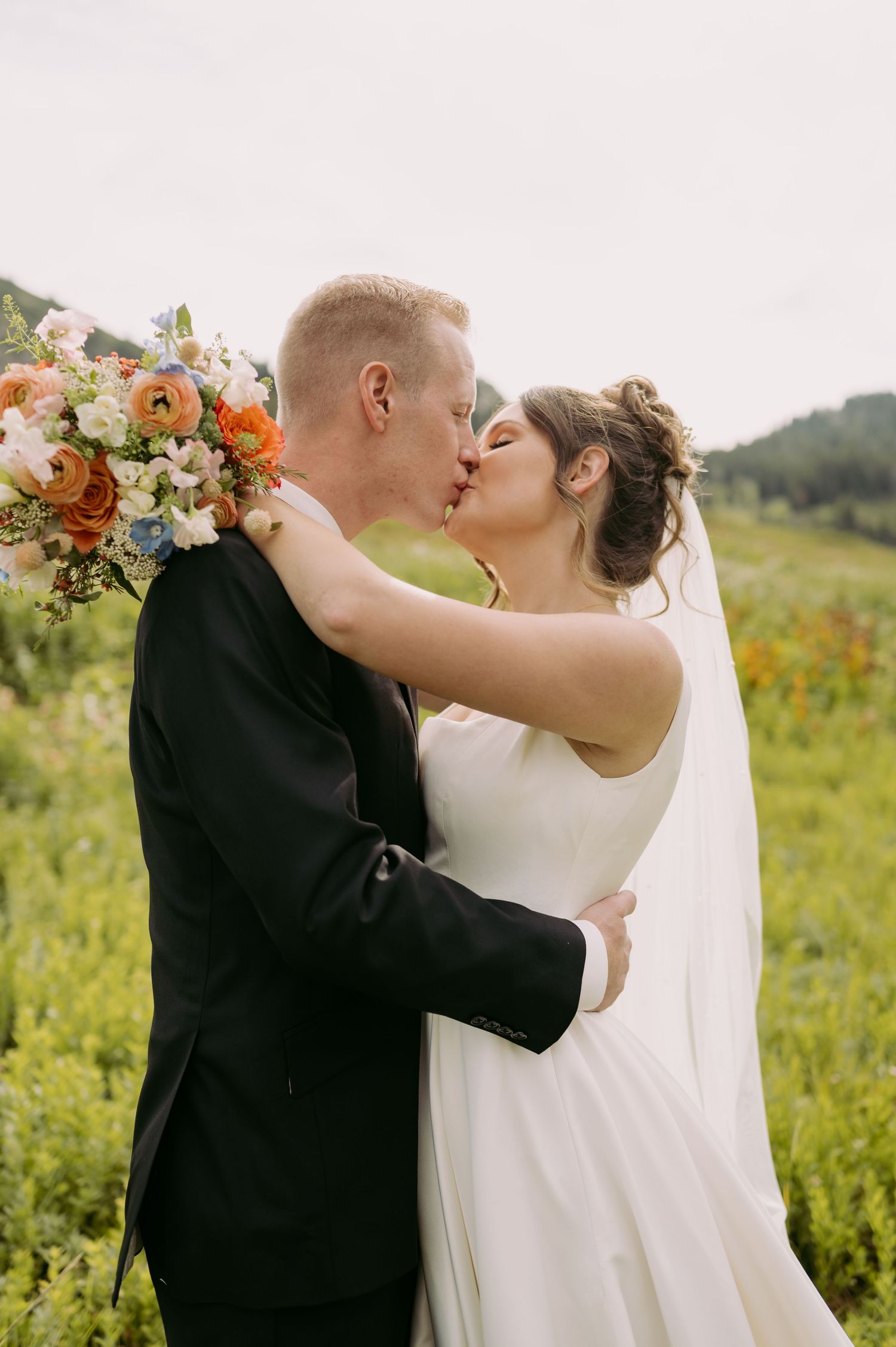 Bride and groom share passionate kiss with colorful bouquet in mountain meadow.