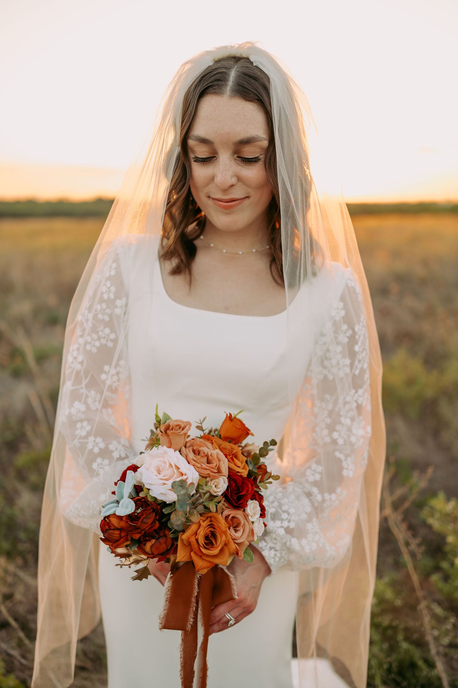 A bride holds a vibrant orange and pink bouquet while wearing a lace-sleeved dress in golden sunlight.