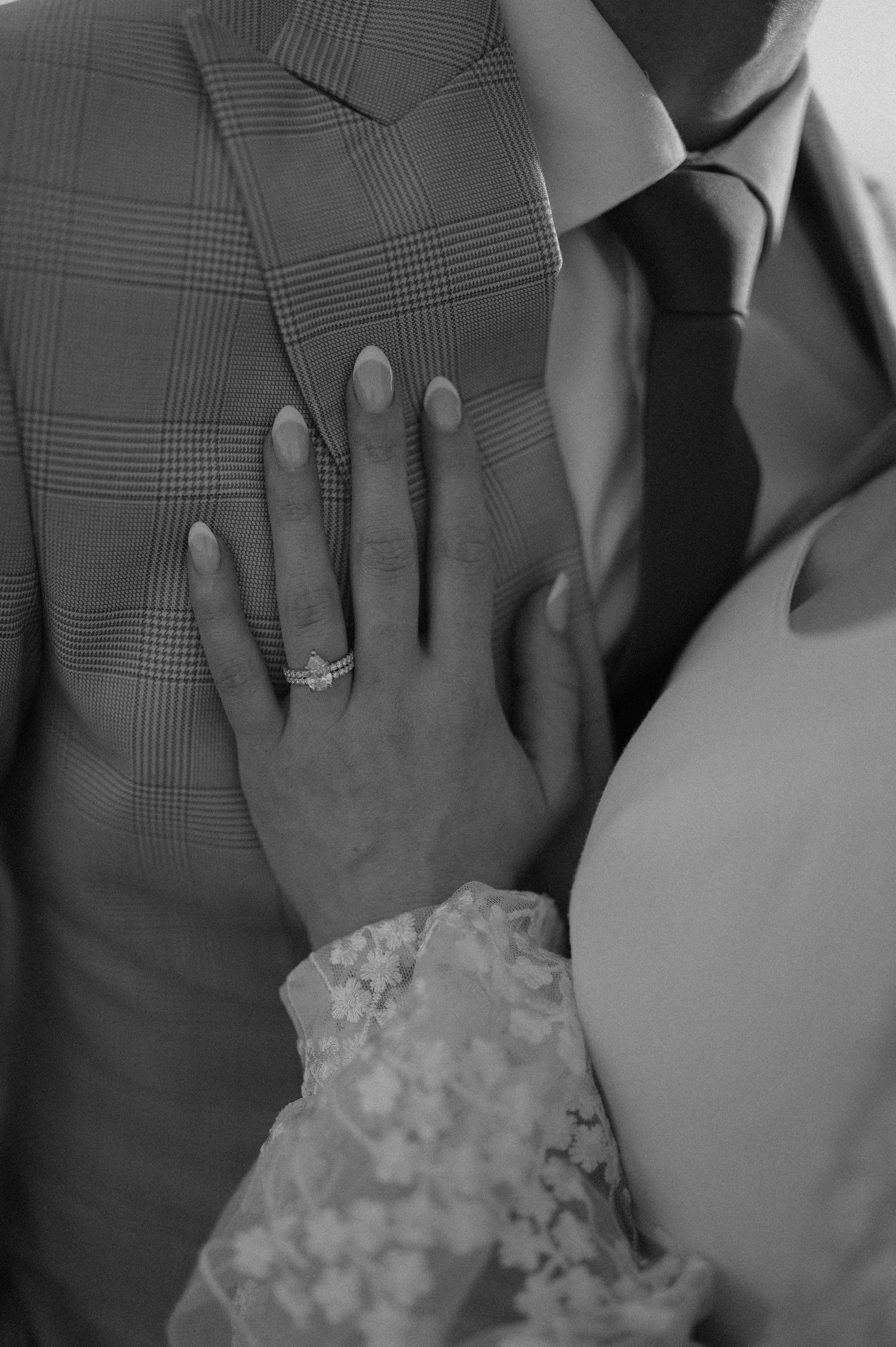 A close-up black and white shot of hands showing wedding rings and lace dress details.
