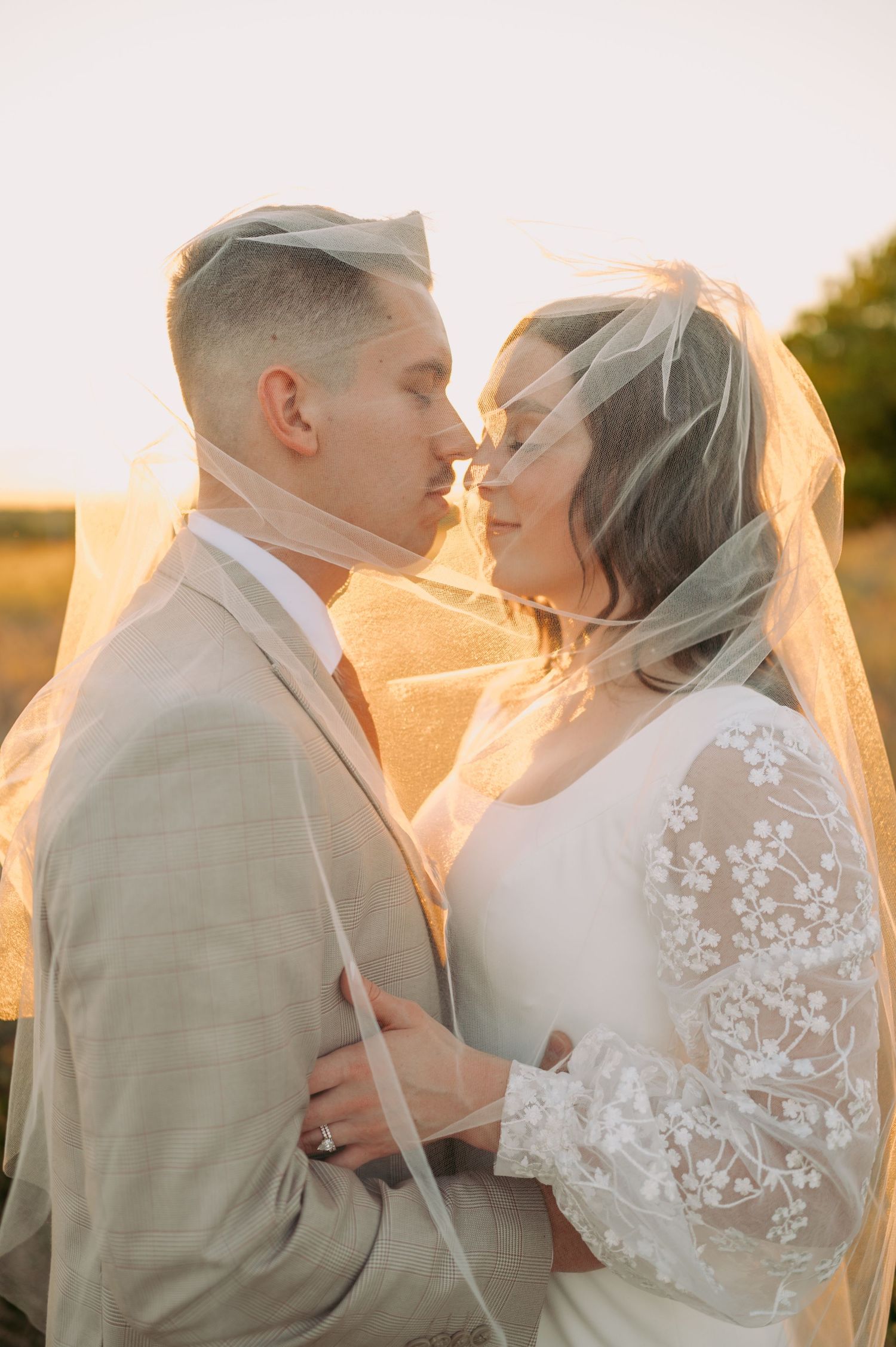 A romantic sunset portrait of newlyweds embracing while wearing light-colored wedding attire.