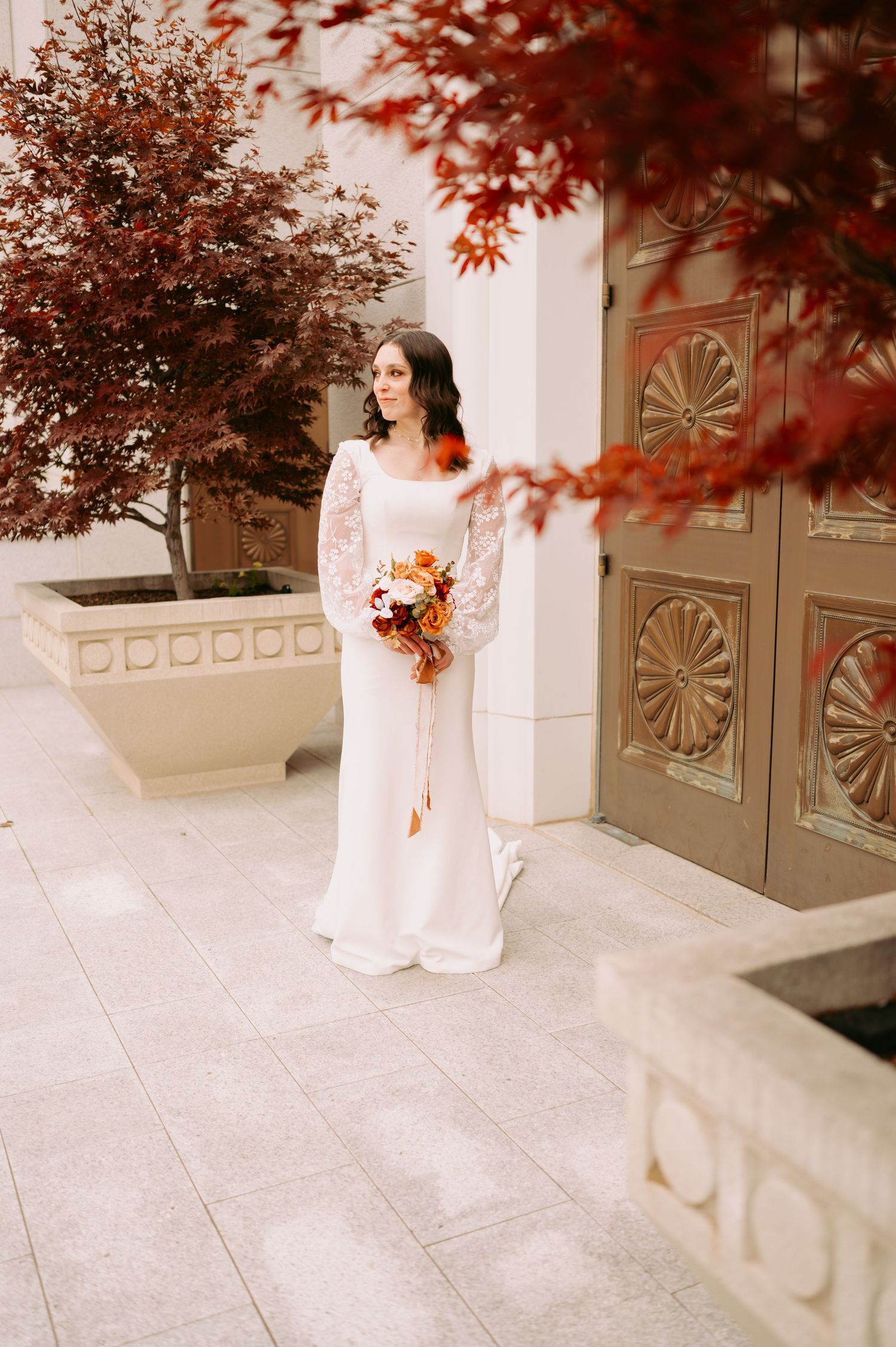 A bride in a lace-sleeved gown poses with autumn flowers against a white temple wall.