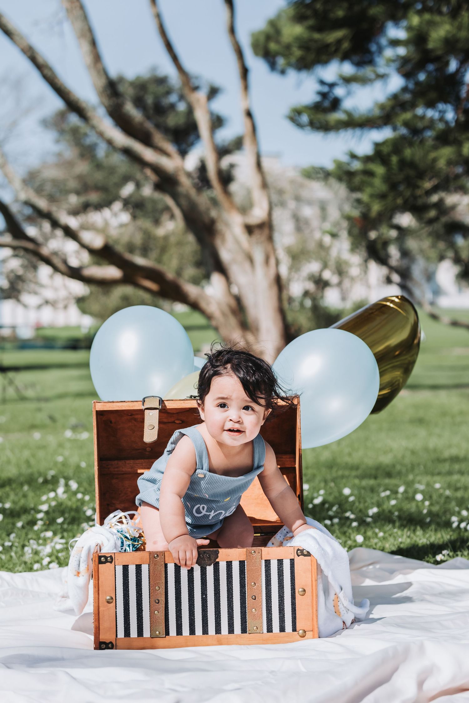 A toddler sits in a vintage wooden chest with blue balloons in a sunny park setting.