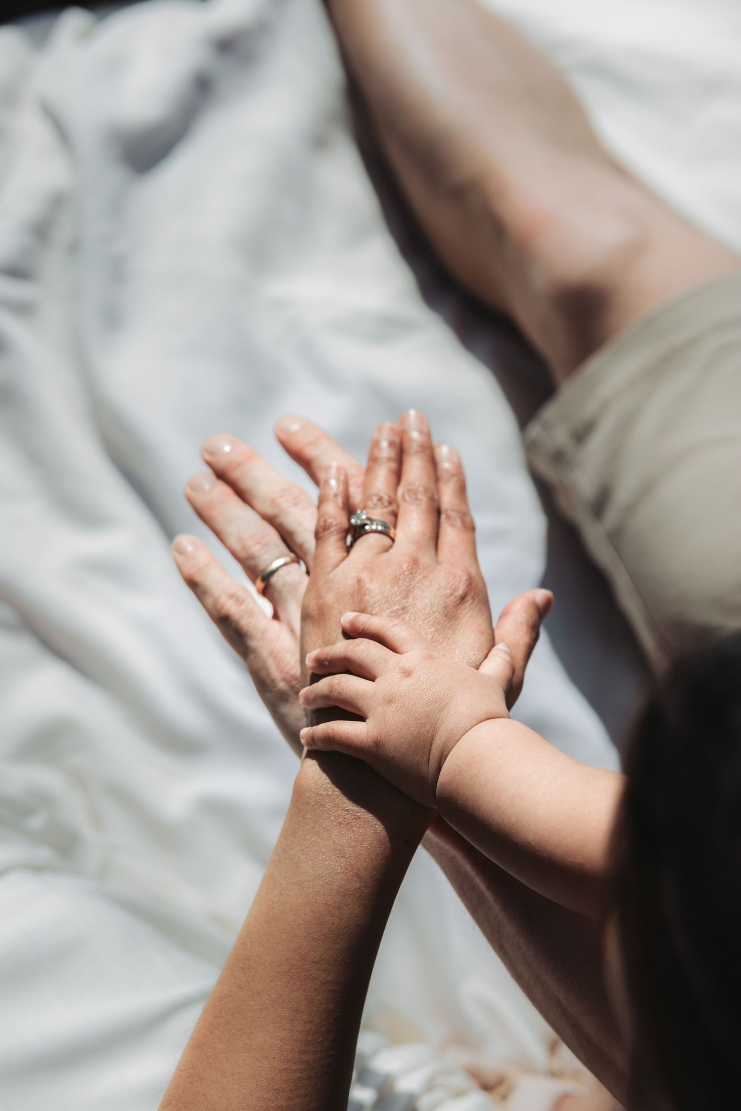 Close-up of adult and child hands touching with wedding rings visible on white fabric.