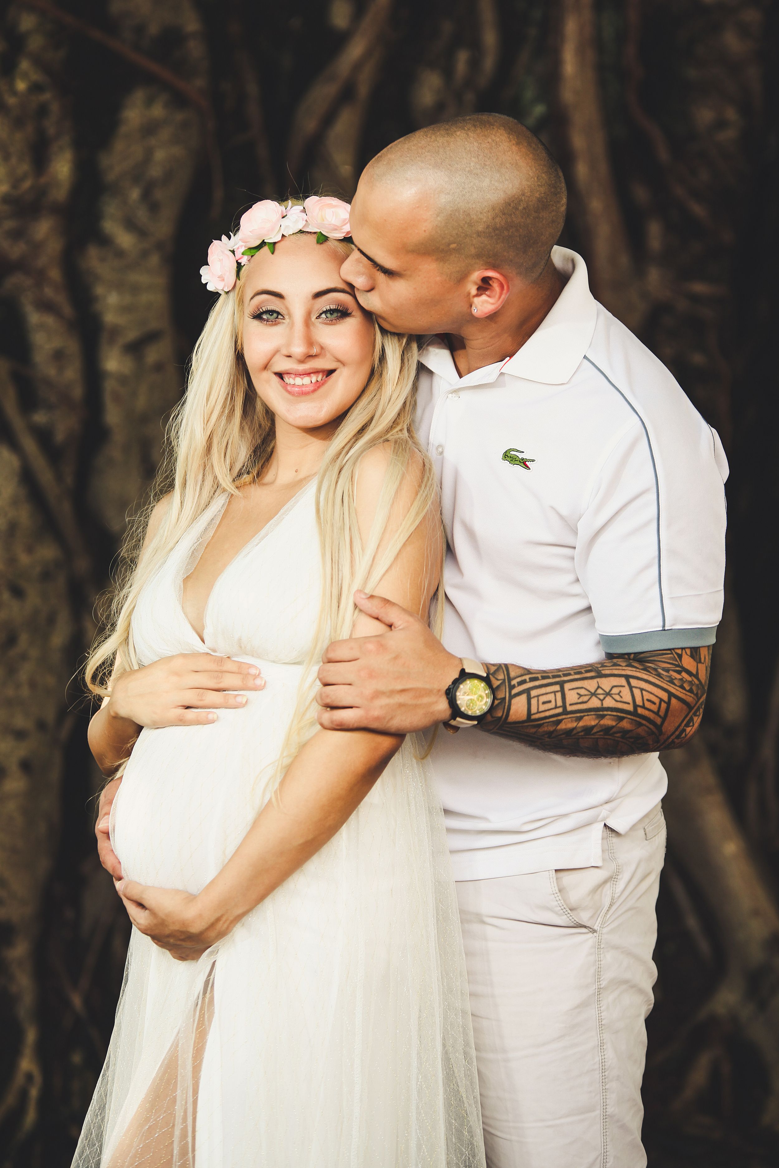 A romantic couple in white outfits embraces during an outdoor photo session against a wooded background.