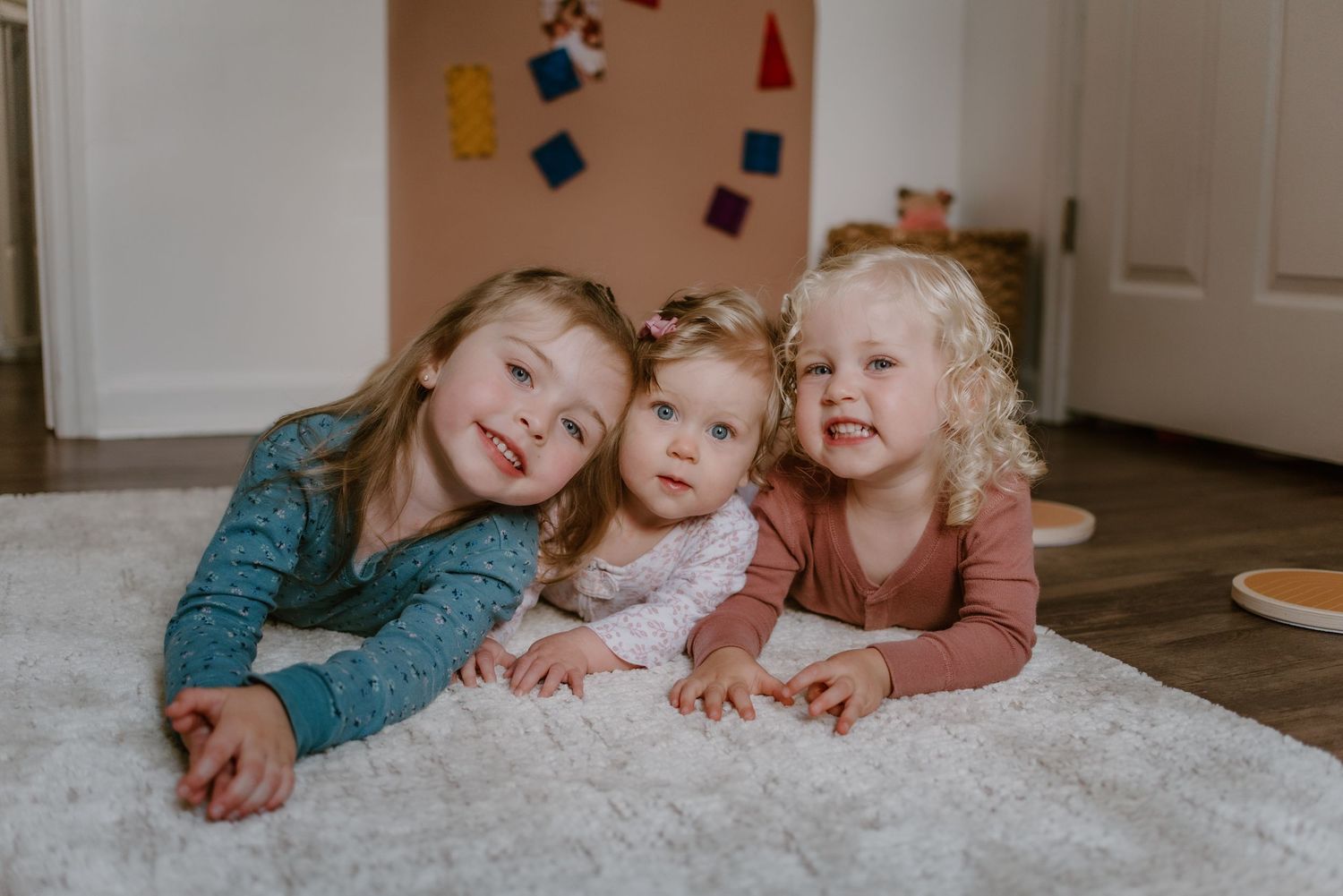 Three children lying together on white carpet while smiling at camera.