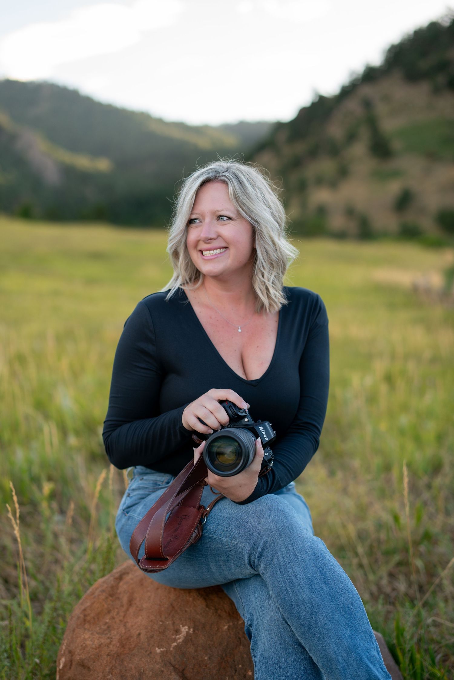 Photographer sitting on rock in mountain meadow holding camera during golden hour.